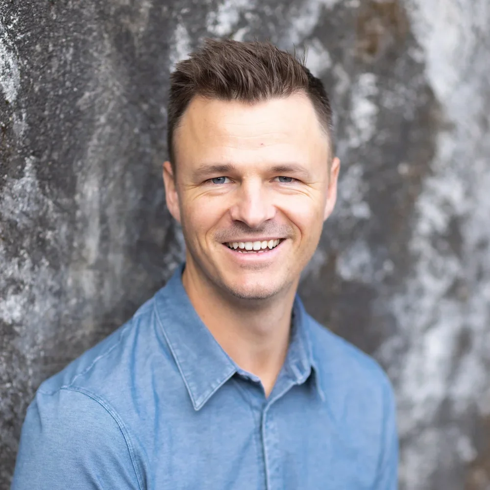 Teacher Sandy at Youngset Preschool appears as a smiling man with short dark hair wearing a blue denim shirt, standing outdoors against a textured gray stone wall.