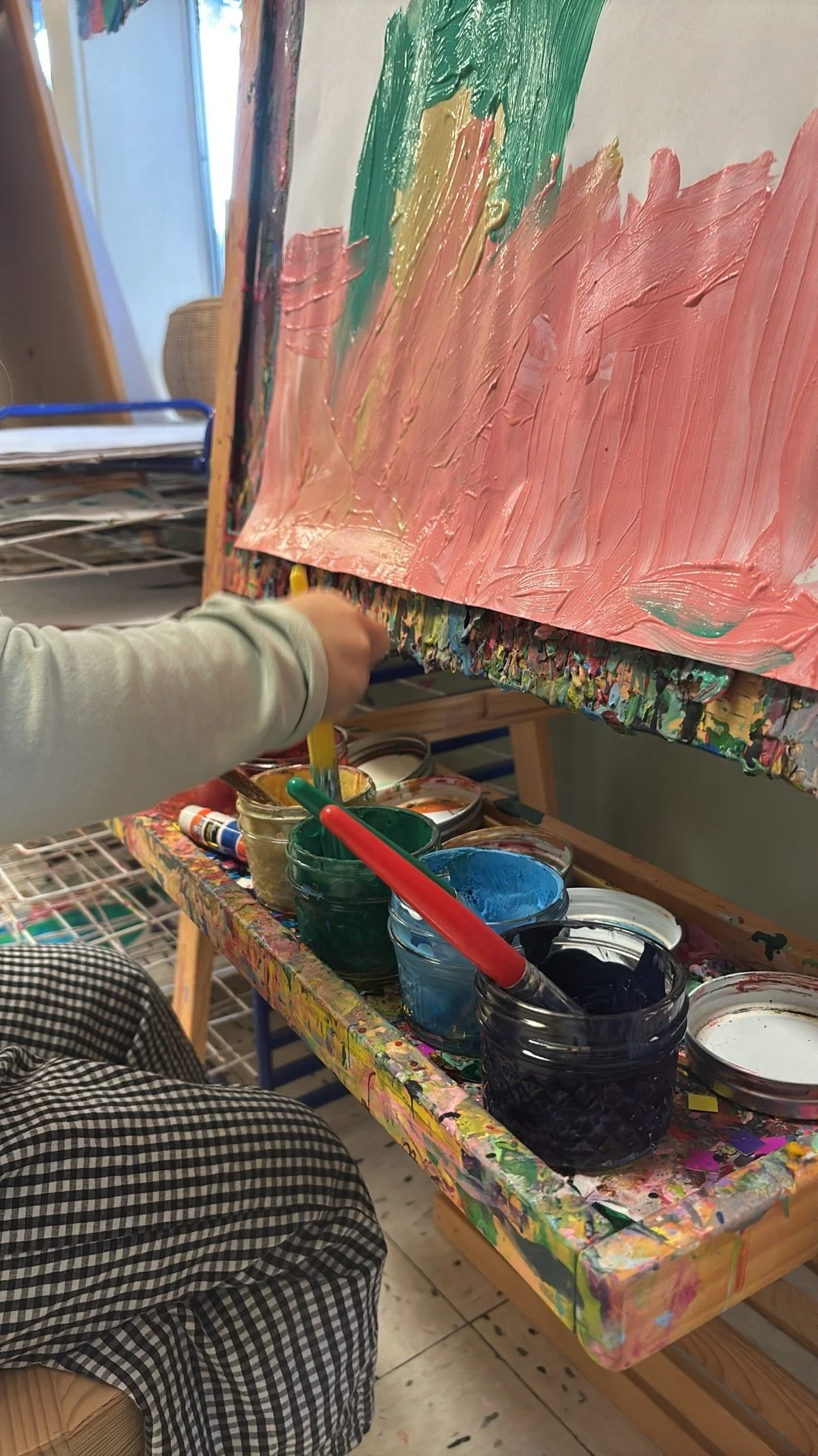 Child at Youngset Preschool painting on a canvas with pink, green, gray, and yellow paint, sitting at a colorful easel with cups of paint and brushes nearby.