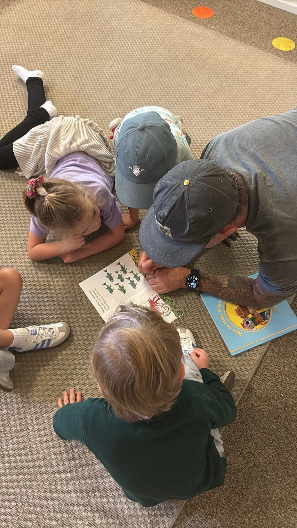 A father volunteering at Youngset Preschool reads a children's book to a group of kids sitting and lying on a carpeted floor.