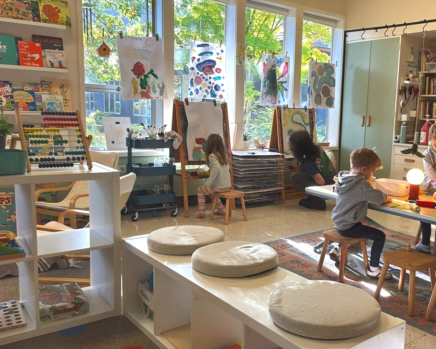 Children and a teacher working on art projects in a brightly lit classroom with large windows and colorful artwork on display.