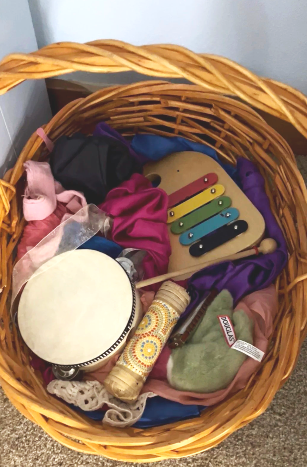 A wicker basket containing a wooden musical shaker, a colorful wooden xylophone, a drum, a wooden hammer, and various cloth items, possibly for children's play or musical activities.