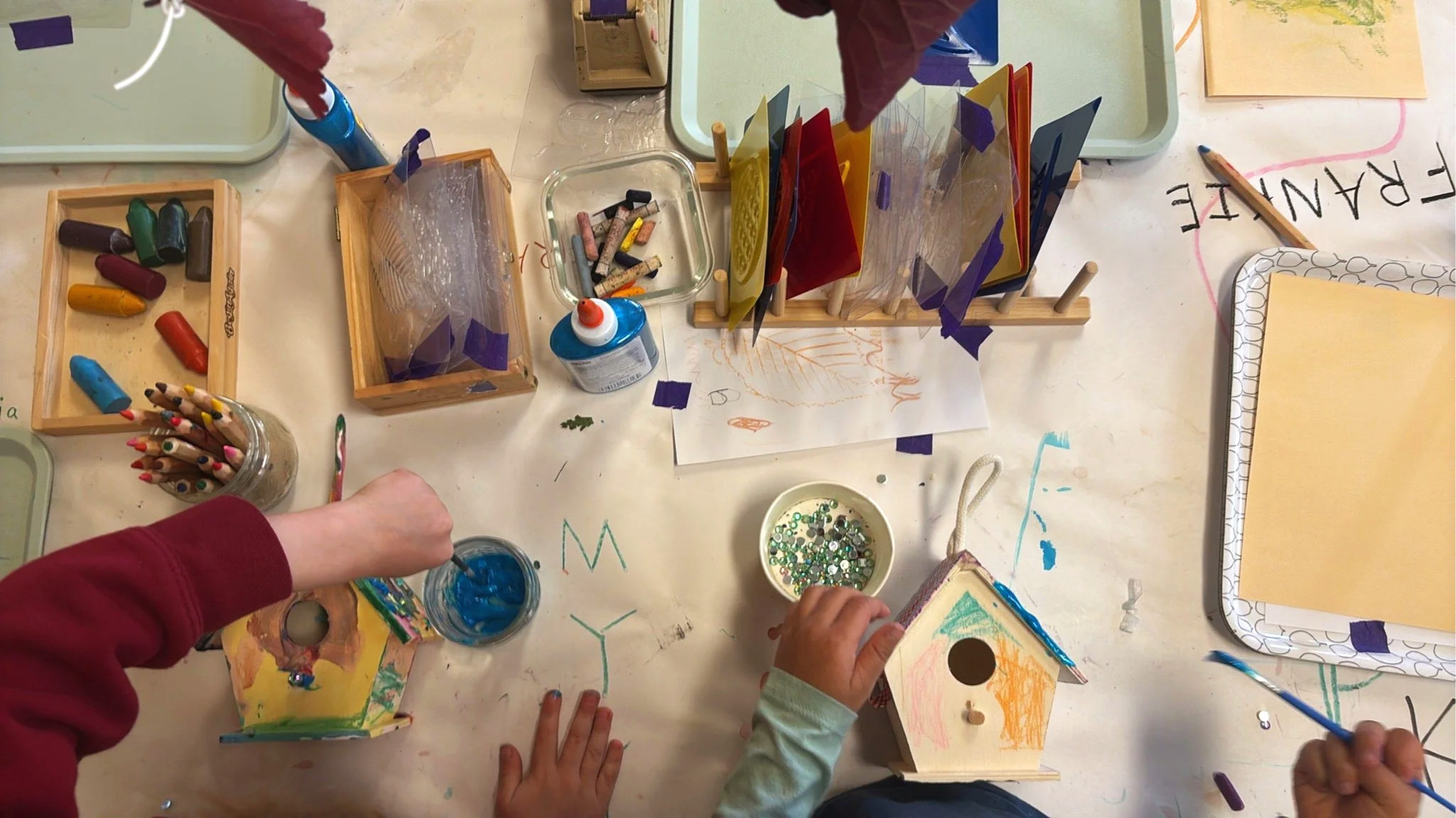 Children's hands and arms are visible at a table engaging in arts and crafts. Various supplies such as colored chalks, drawing paper, sequins, glue, and decorative items are scattered across the table. The surface has handwritten words and colorful markings, indicating an active creative activity.
