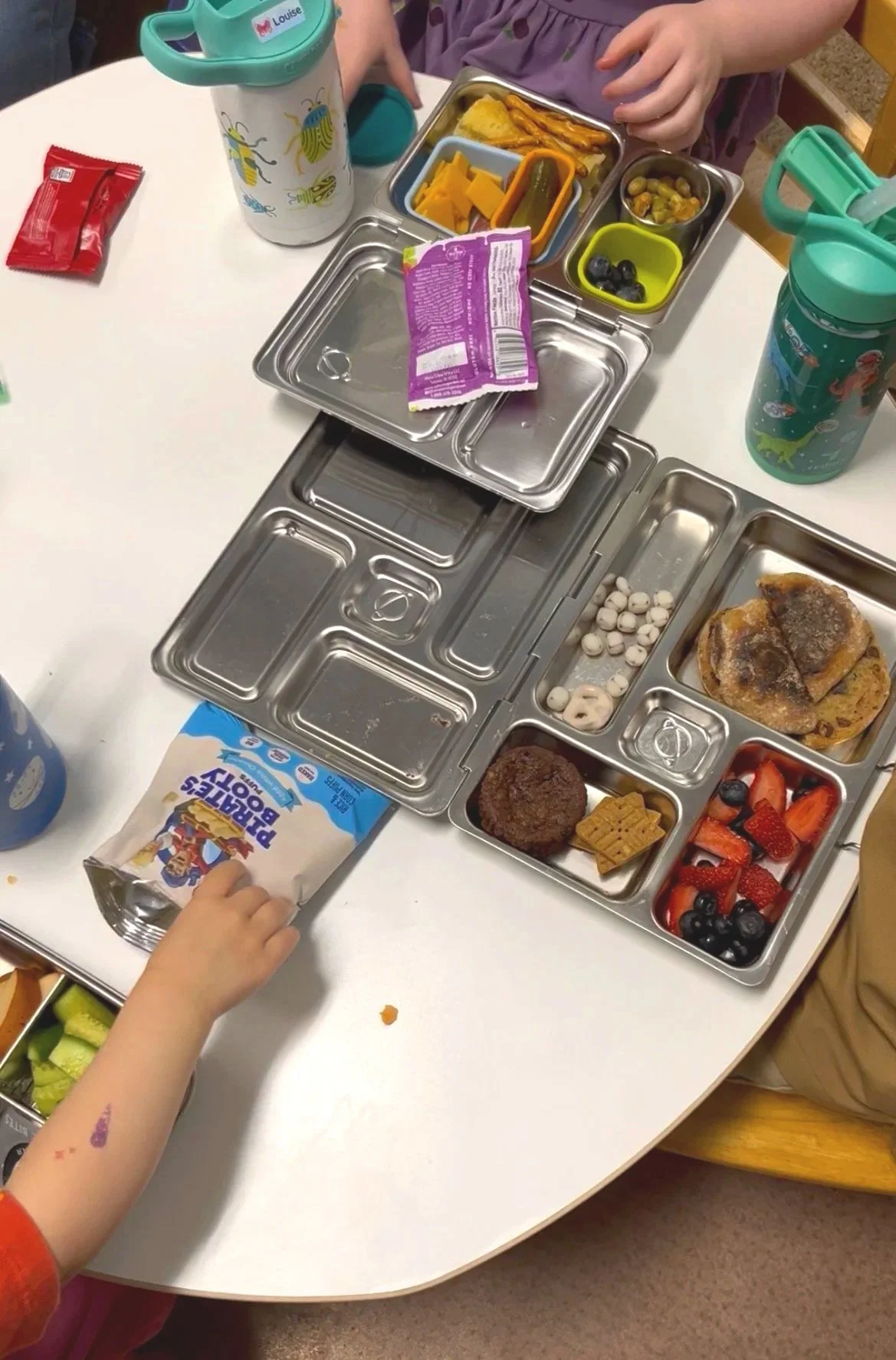 A children's lunchbox with various compartments filled with food, including strawberries, blueberries, crackers, a meat patty, a piece of cooked chicken, vegetables, and cheese, placed on a white table alongside a water bottle, a juice box, and other snacks, with children nearby.