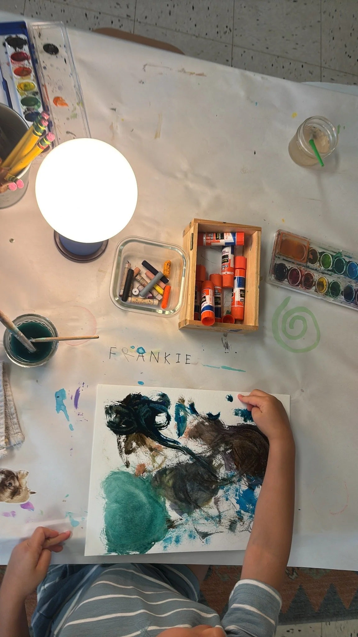 A child at Youngset Preschool works on a colorful abstract painting on a sheet of paper, surrounded by art supplies such as glue sticks, watercolor paints, brushes, and a lamp on a worktable.