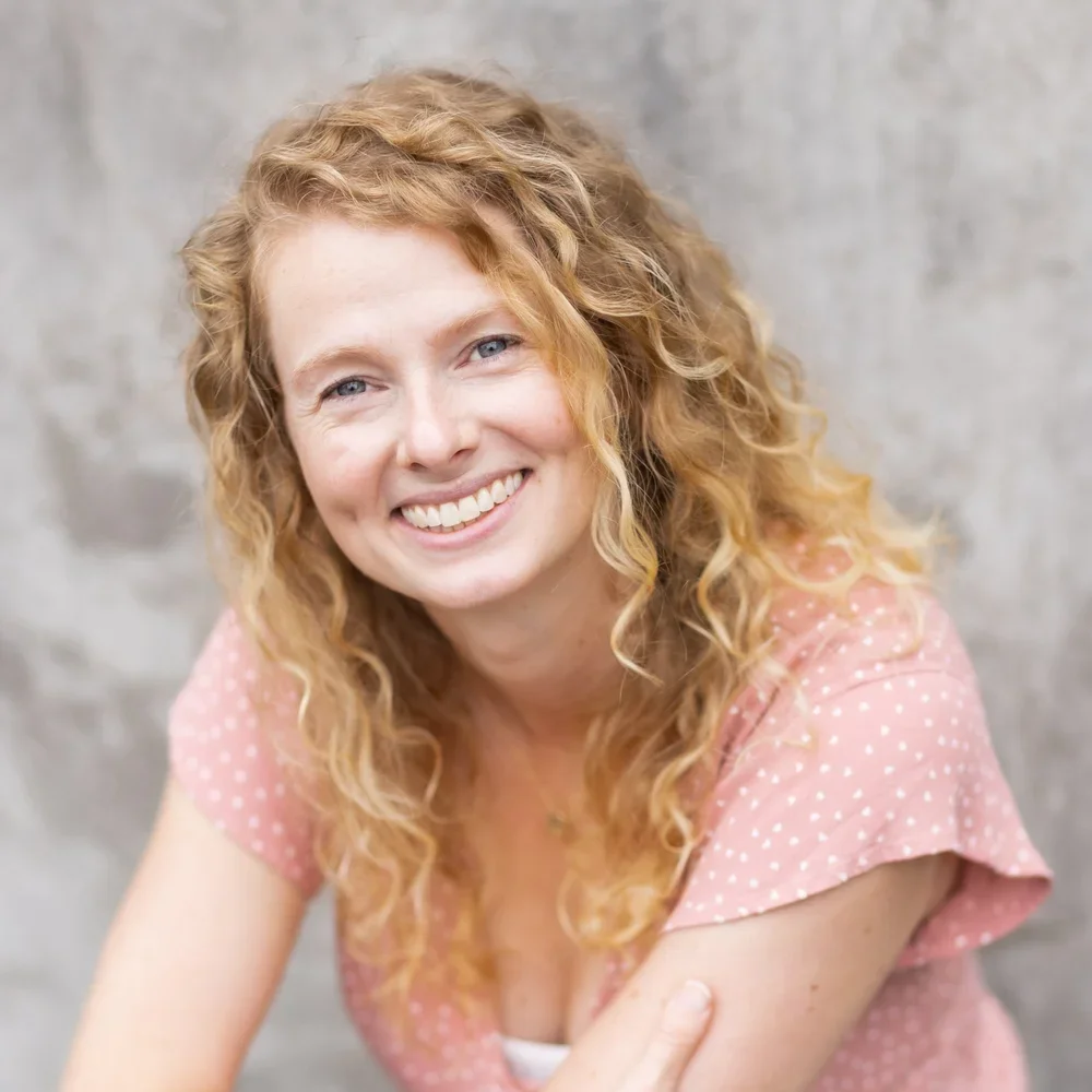 Teacher Emily at Youngset Preschool appears as a woman with curly red hair smiling in front of a gray stone wall.