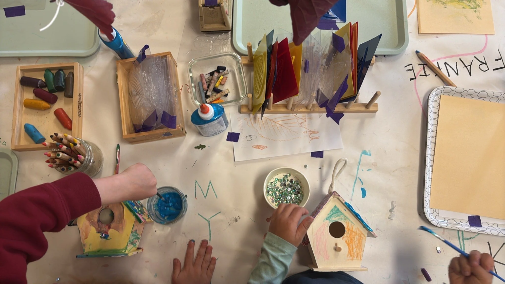 Children and adults engaged in arts and crafts activities at a table filled with colored chalks, paper, glue, and decorative items like sequins and colored acrylic sheets at Youngset Preschool.