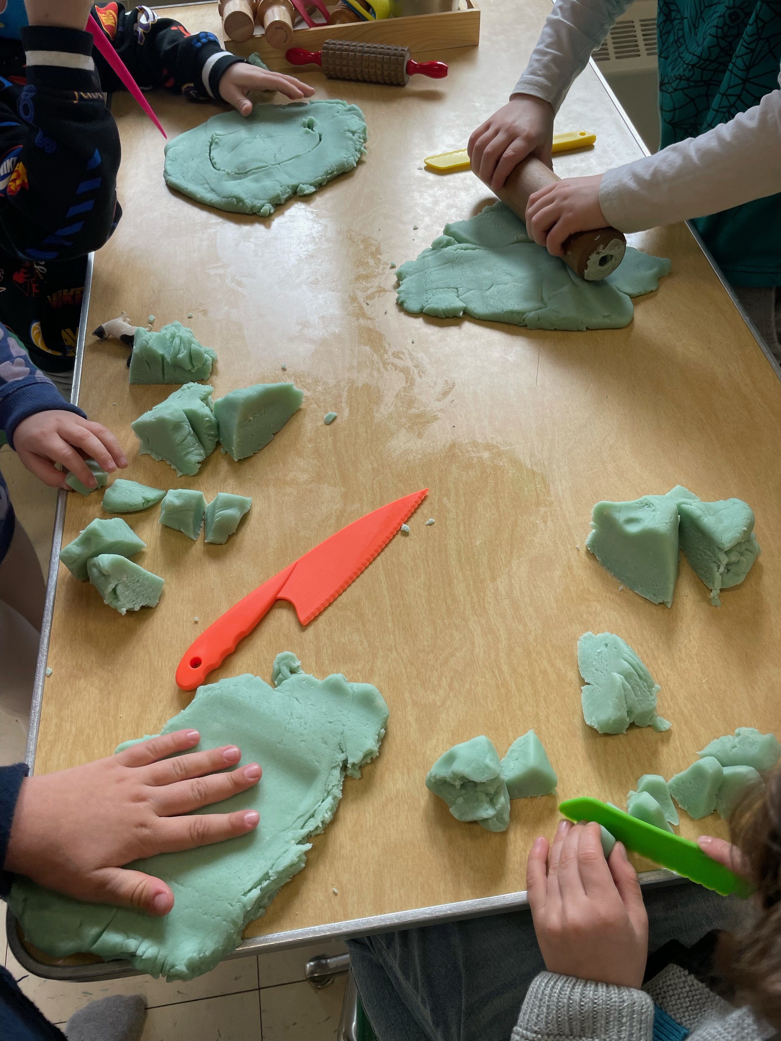 Children at Youngset Preschool playing with green modeling clay on a wooden table, using their hands and a rolling pin to mold and cut the clay.