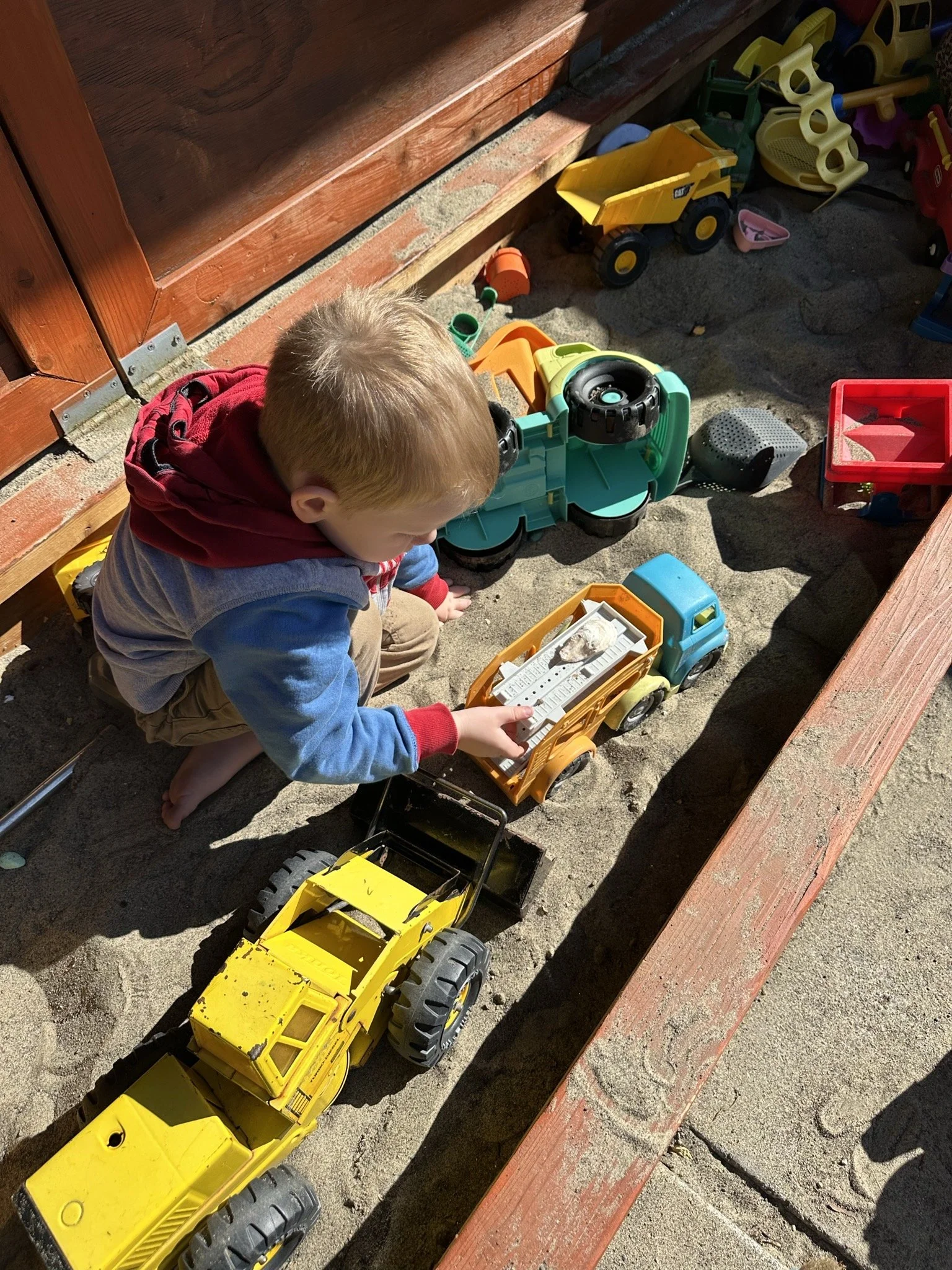 A young boy at Youngset Preschool playing in sand with toy trucks and construction toys at a sandbox area.