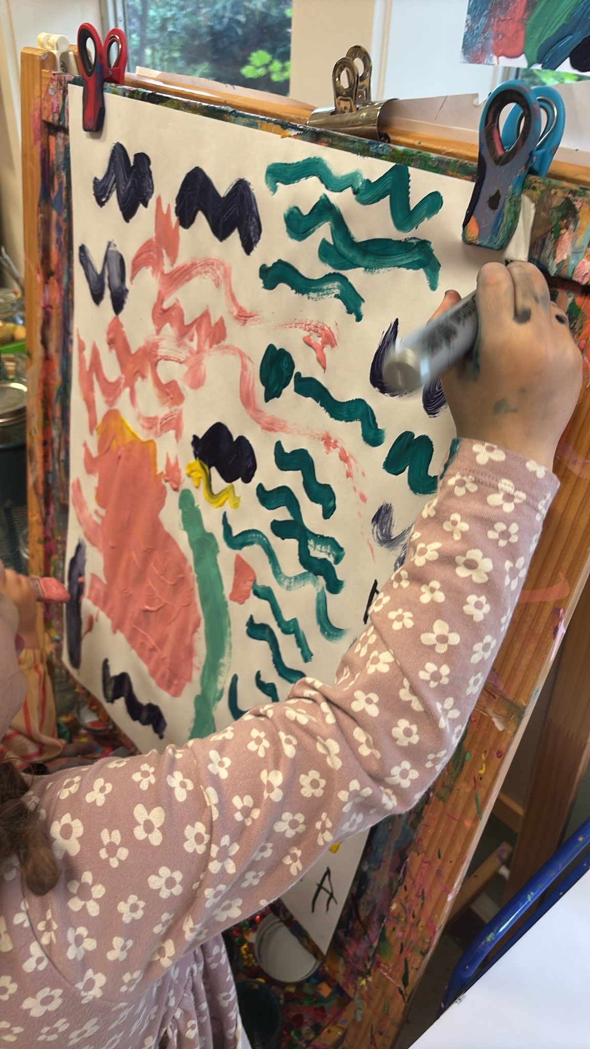 Child at Youngset Preschool wearing a pink dress with white flowers painting on a large sheet of paper attached to an easel with colorful clothespins. The child is using a white paint marker and has paint on their hand.