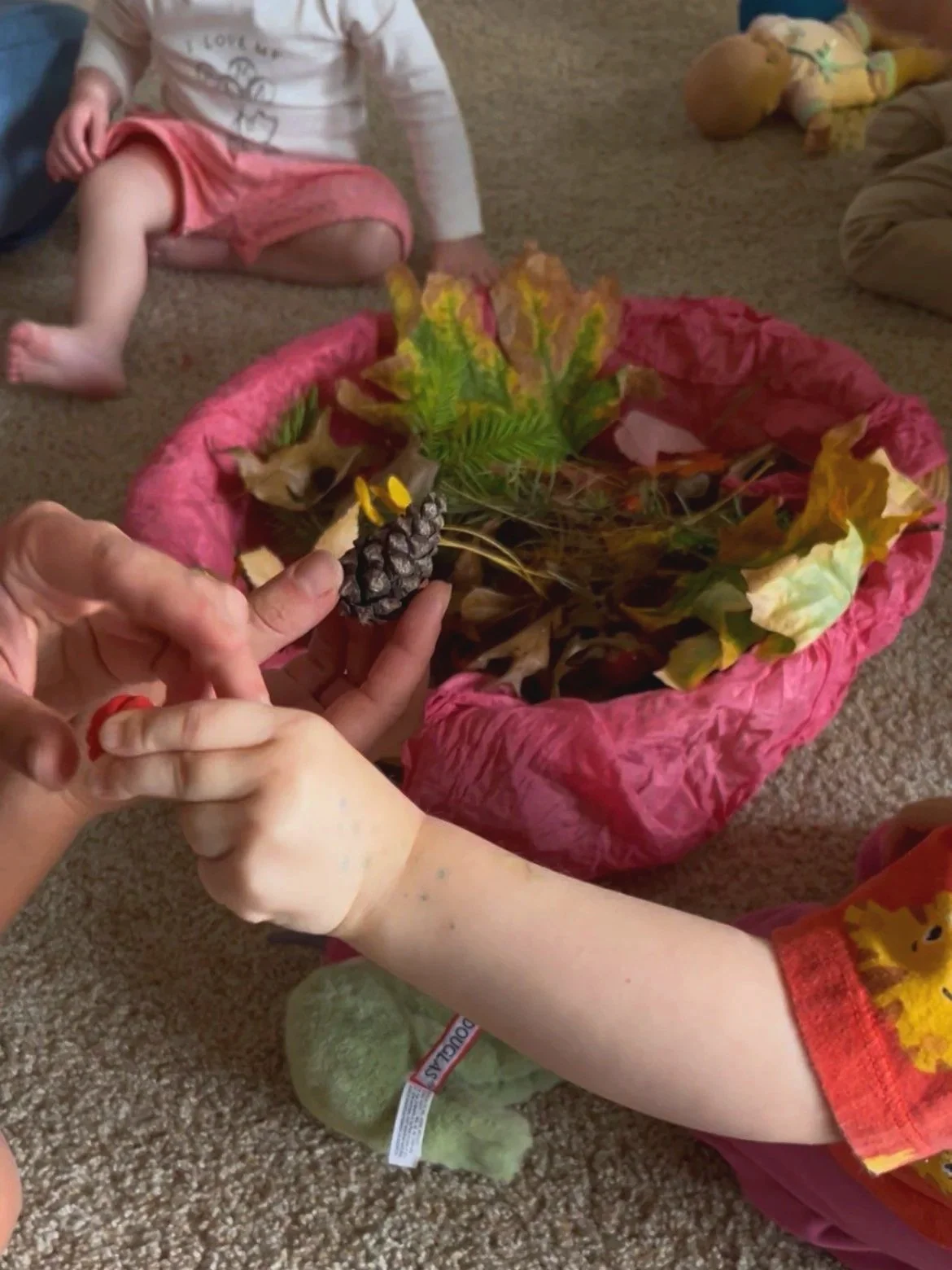 Children's hands making a craft with a pine cone, leaves, and twigs in a pink basket, while other children sit on the carpet in the background.