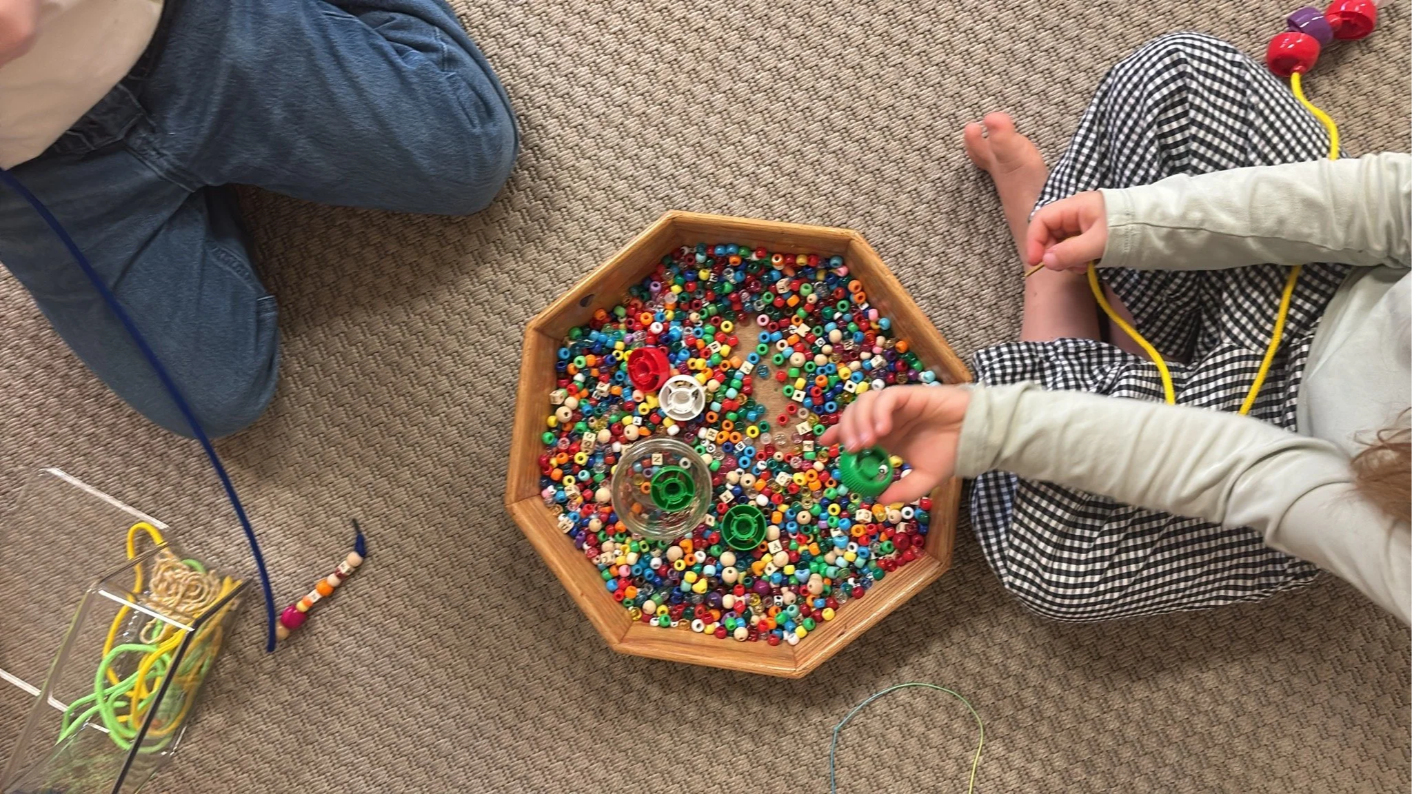 Children playing with colorful beads and crafting supplies on a beige carpeted floor.