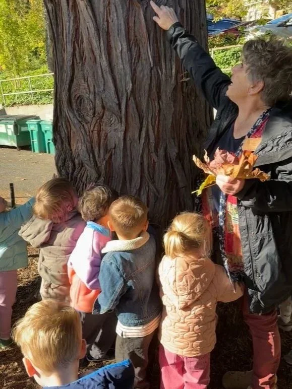 A group of young children gathered around and touching a large tree trunk while an adult woman explains something about it.