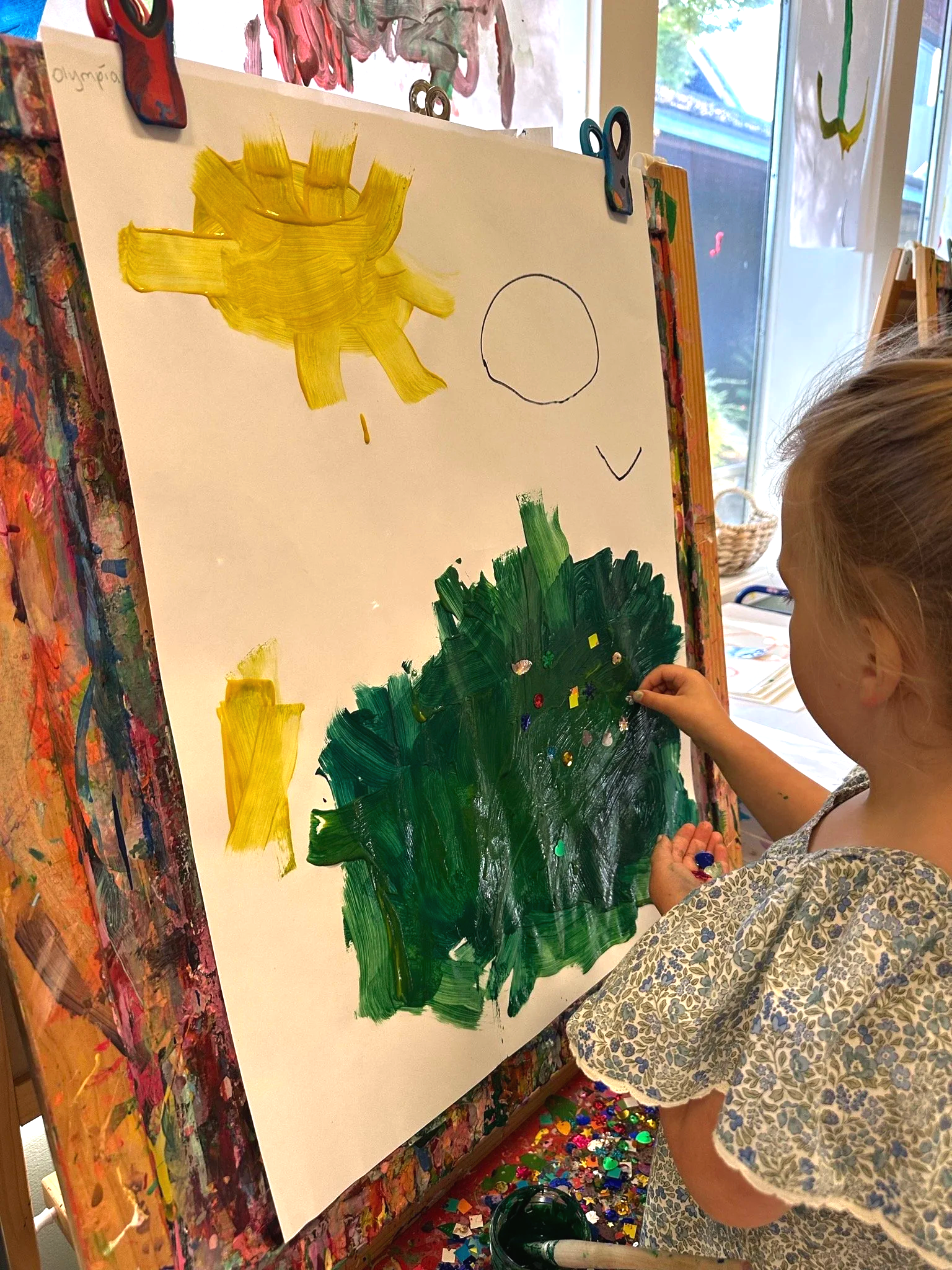 A young girl paints a yellow sun and a green tree on a large easel at Youngset Preschool using bright yellow and green paint, with various colors of glitter on the bottom of the canvas, in a classroom or art studio setting.