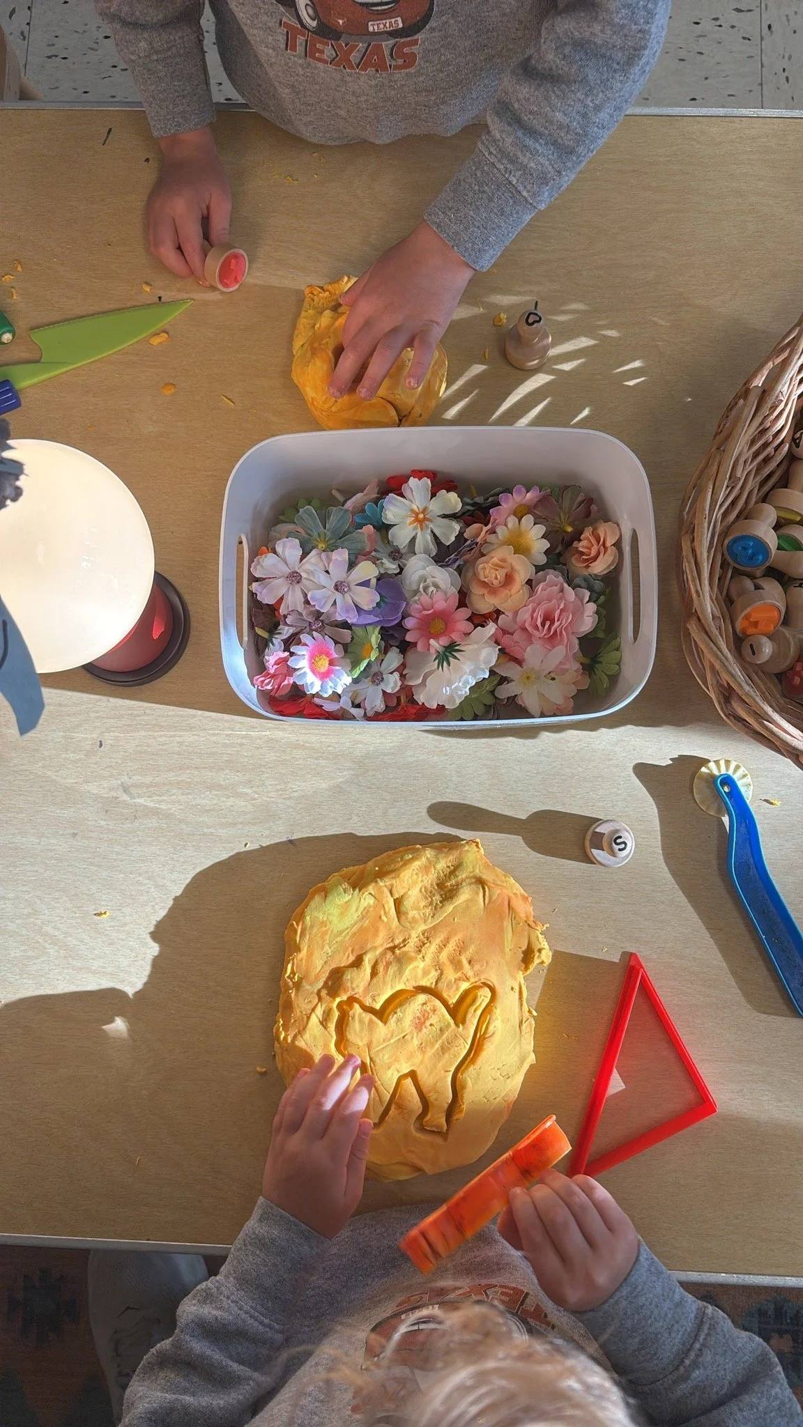 Children decorating and making indentions on play-doh on a table, with various carving tools, a basket of artificial flowers, and a lamp nearby.