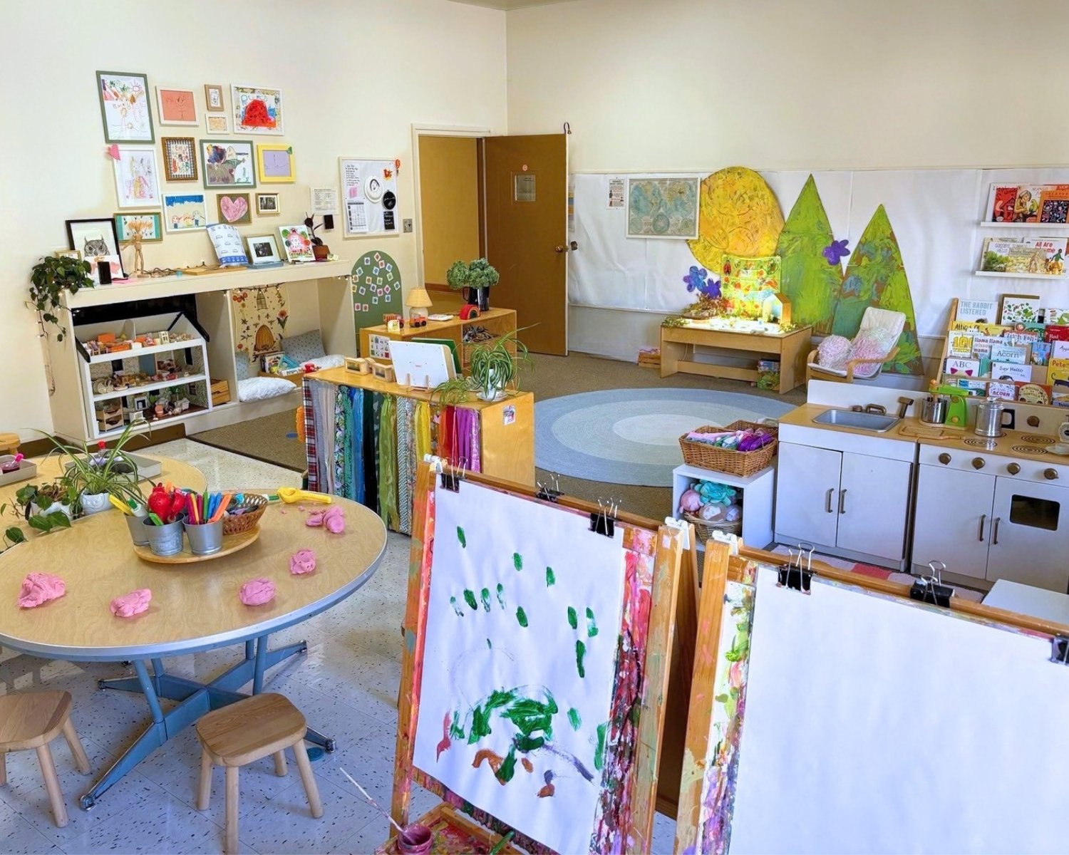 A colorful preschool classroom at Youngset Preschool with children's artwork on the wall, small furniture, art supplies, and play areas, including a reading nook and a pretend kitchen.