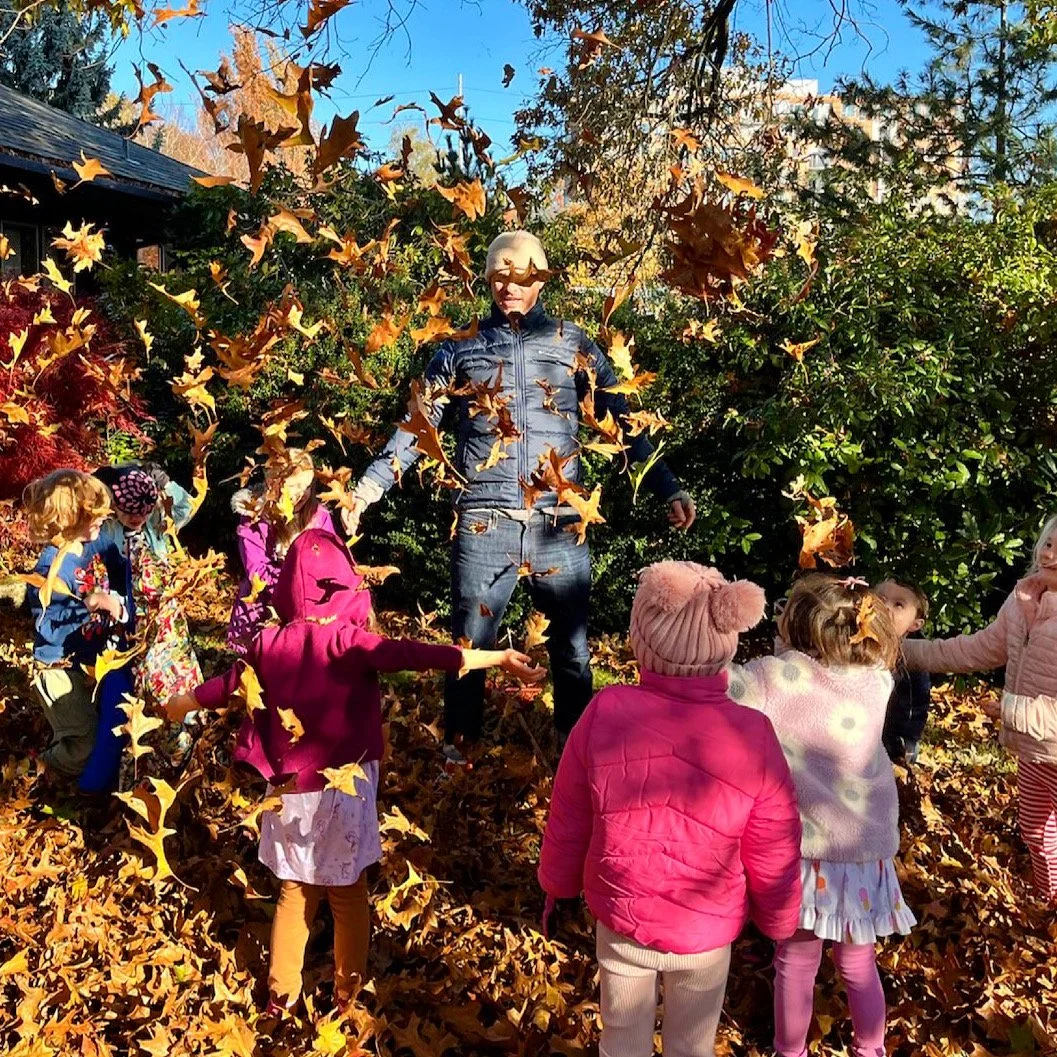 Teacher Sandy standing on a pile of leaves surrounded by children playing in the outside courtyard at Youngset Preschool during fall.