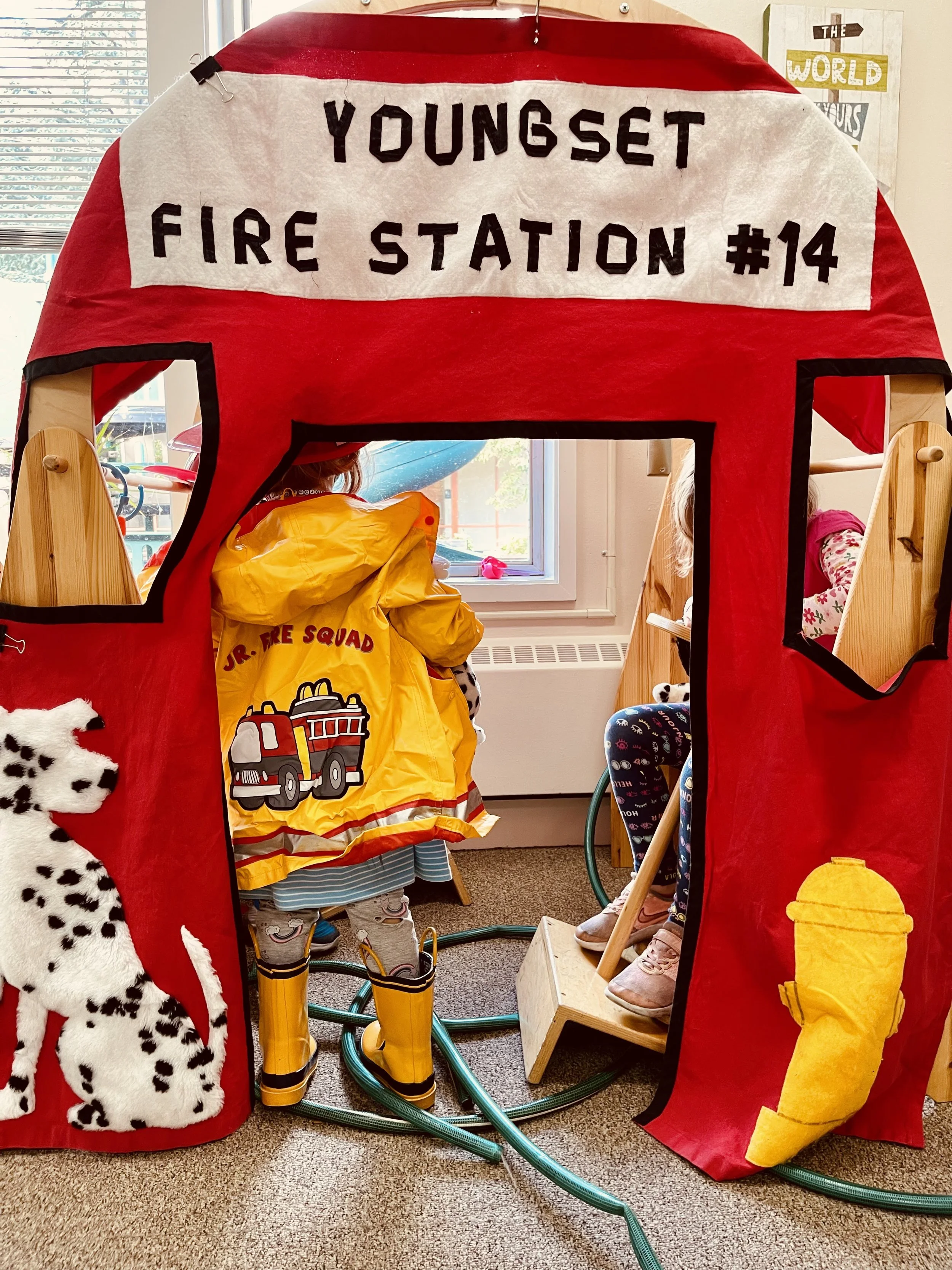 Children playing inside a small red playhouse at Youngset Preschool styled as a fire station with a sign that reads "Youngset Fire Station #14." The playhouse has windows and a door, with children inside, some wearing firefighter costumes and boots. 