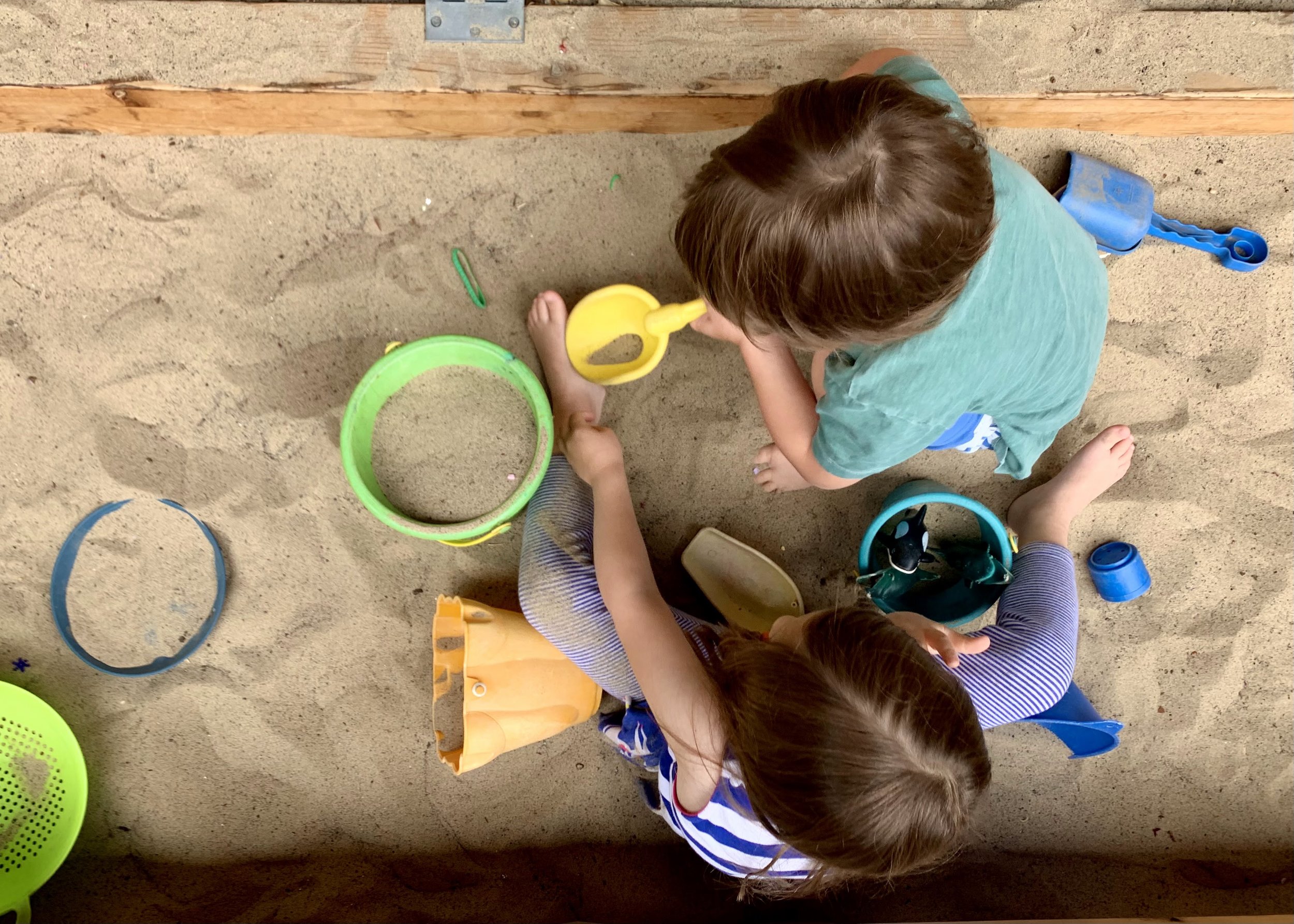 A top-down view of two children playing in a sandbox, surrounded by colorful plastic buckets and toys.