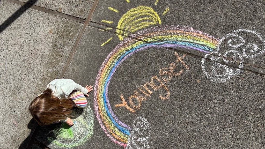 Child drawing with chalk on pavement, creating a rainbow with clouds and the word 'Youngset' in the middle.