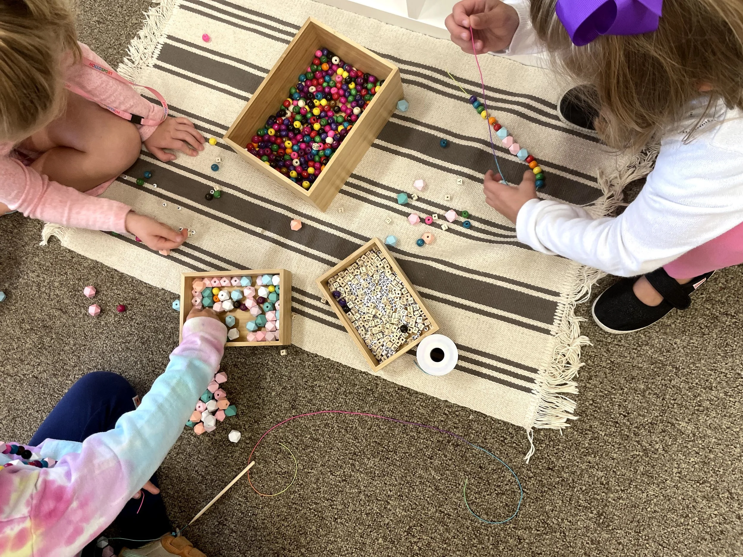 Two children at Youngset Preschool are sitting on a striped rug playing with colorful beads and string to make jewelry. Several containers of beads are scattered on the rug around them.