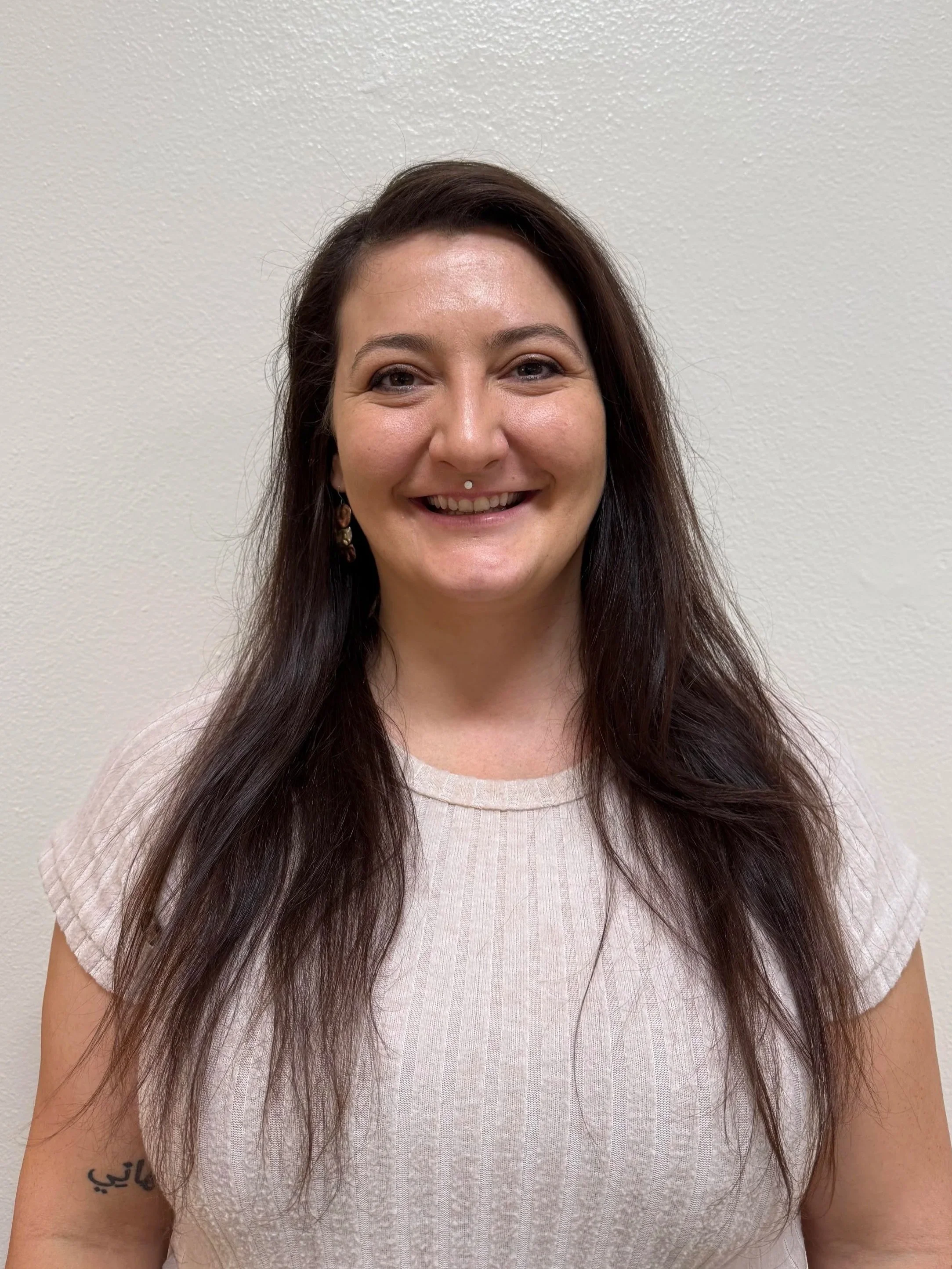 Teacher Laura Youngset Preschool appears as a woman with long brown hair smiling, wearing earrings and a white top, standing against a plain off-white wall.