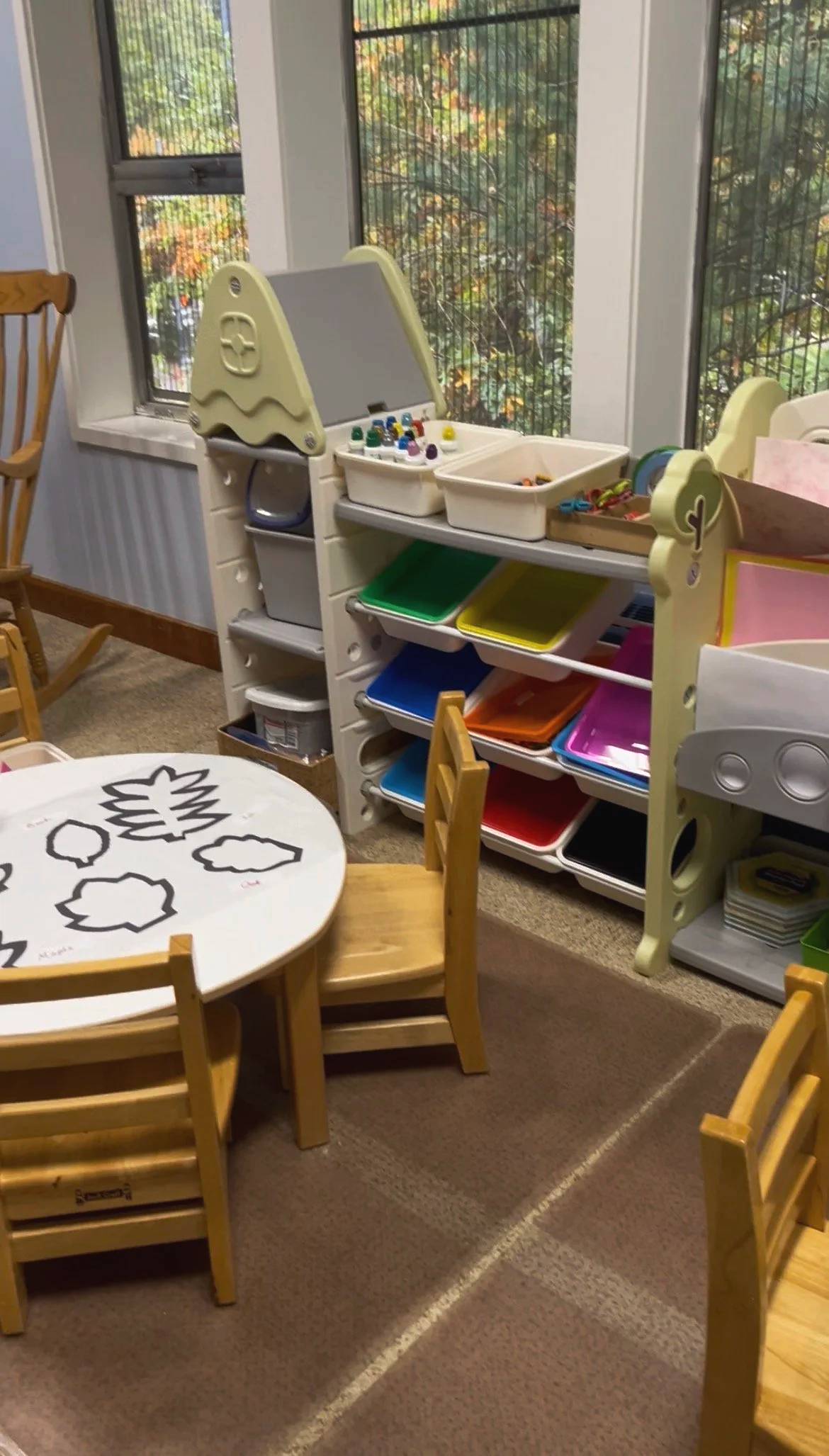 A children's art and play area with a round table and small chairs, a white bookshelf with trays of craft supplies, and large windows showing trees outside.