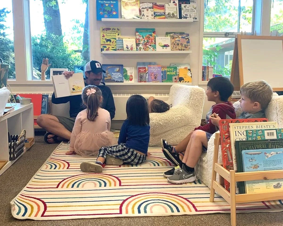 A father volunteering at Youngset Preschool is reading a book to a group of children in a cozy, well-lit corner of the classroom with bookshelves and large windows showing green trees outside.