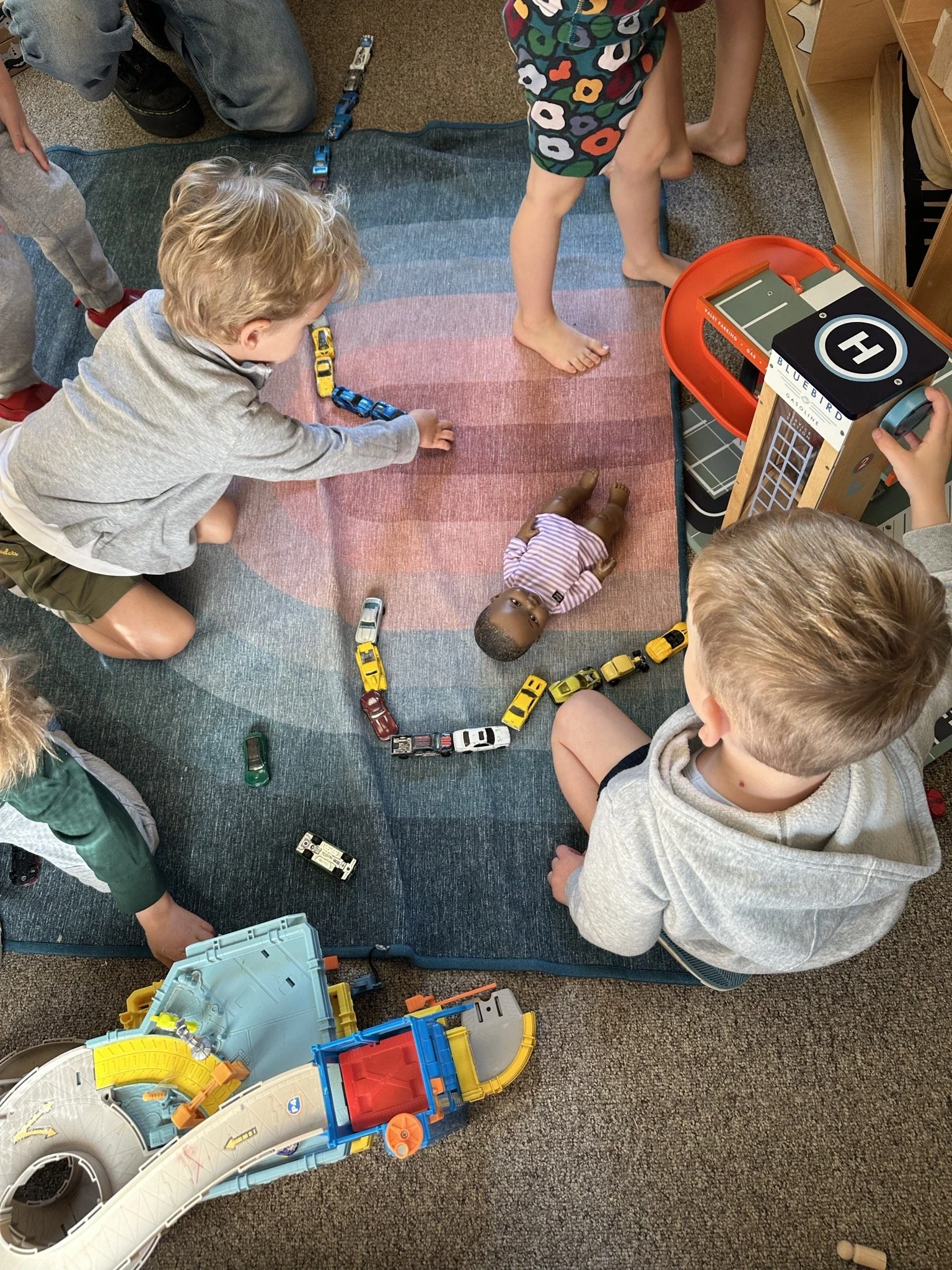 Children playing with a toy race track and cars on a carpeted floor at Youngset Preschool, some children are seated and some are standing around a toy area.