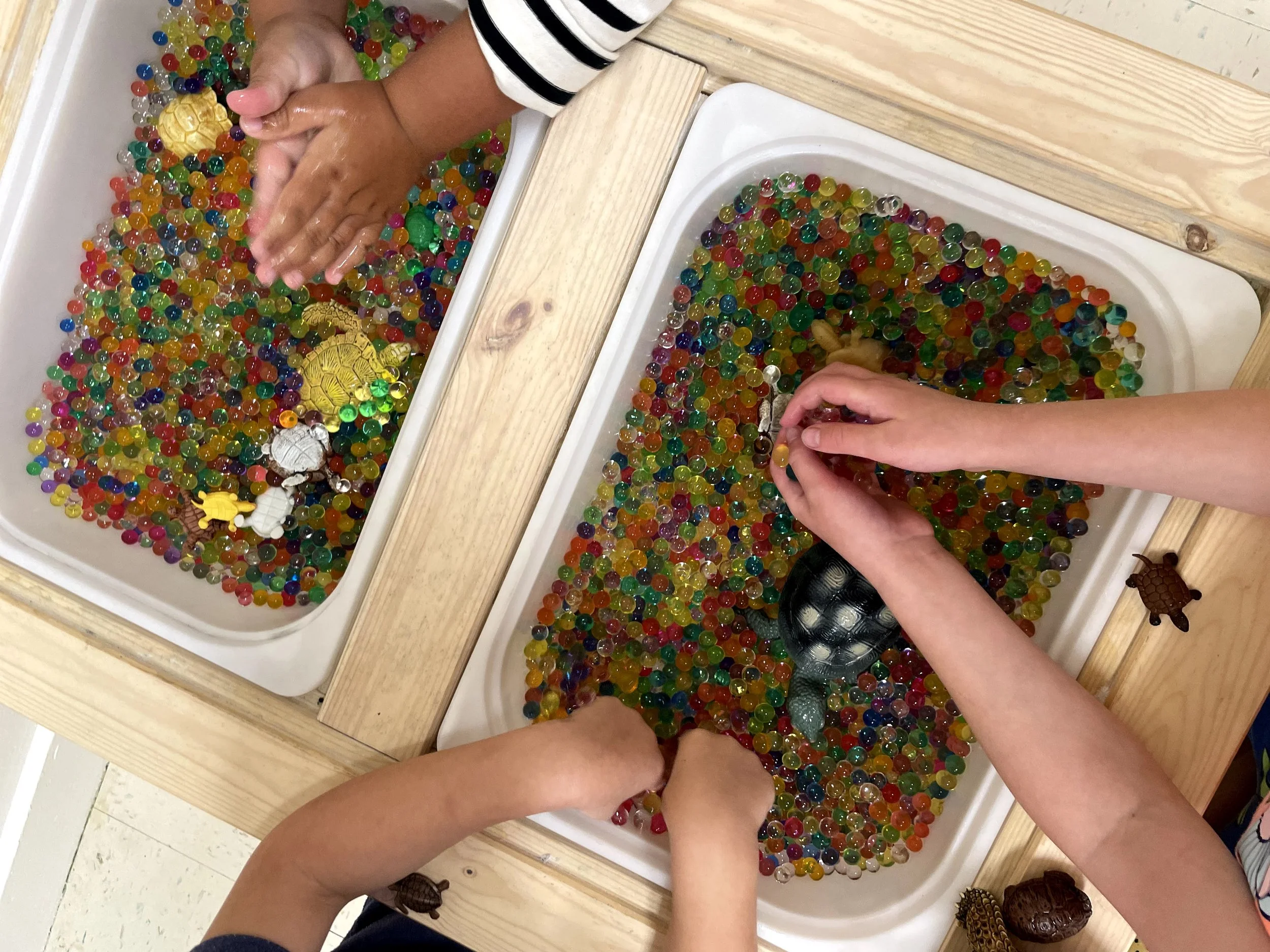 Children playing with colorful water beads in sensory bins, some toy animals in the water, on a wooden table.