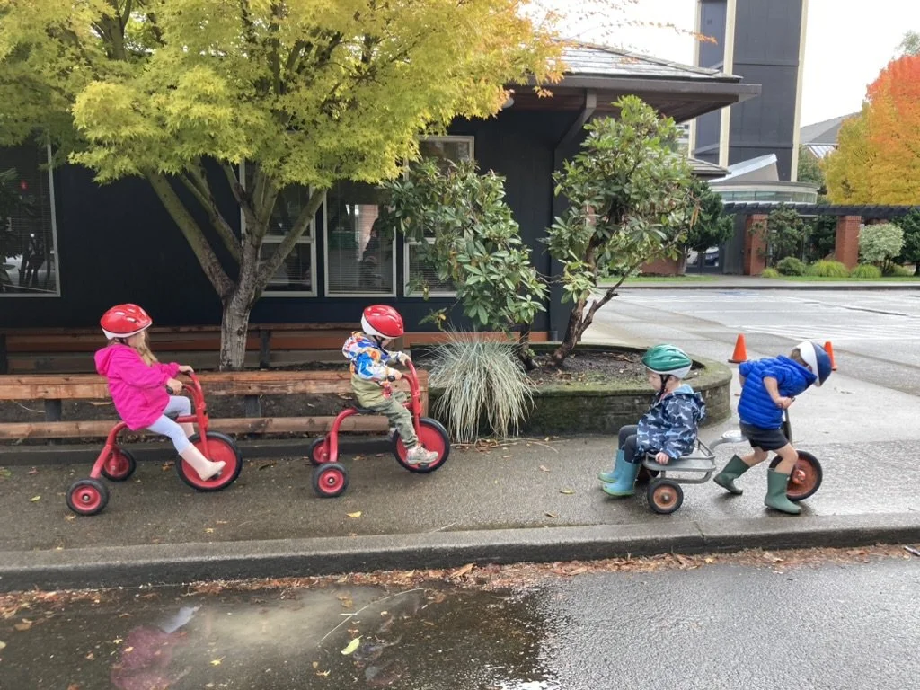 Four children at Youngset Preschool wearing helmets riding tricycles and a small wagon on a sidewalk near a house with trees and bushes.