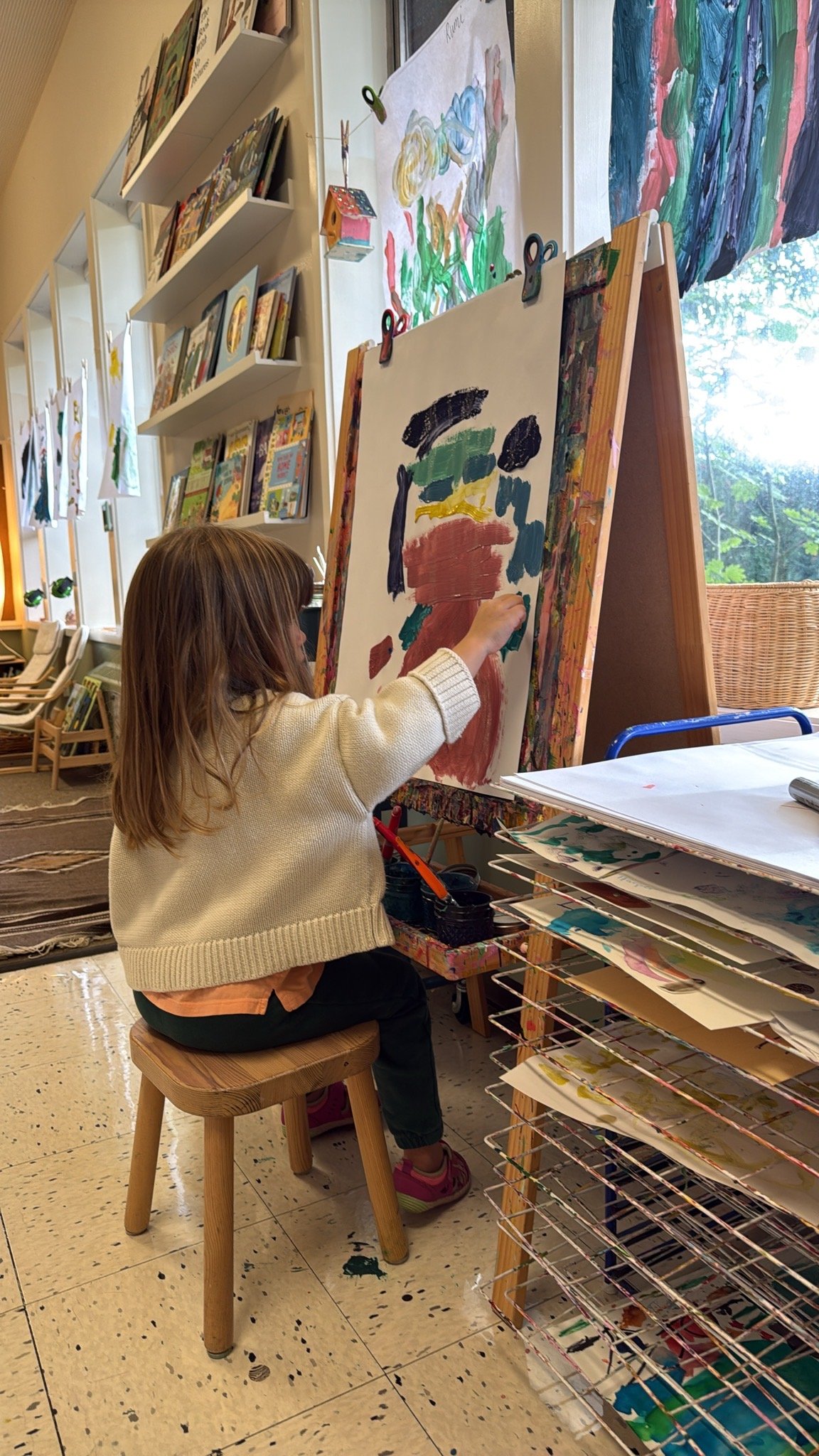 A young girl with brown hair, wearing a white sweater and dark pants, is sitting on a wooden stool while painting on a large canvas at Youngset Preschool. The girl is focused on her painting, which features abstract colorful brushstrokes.