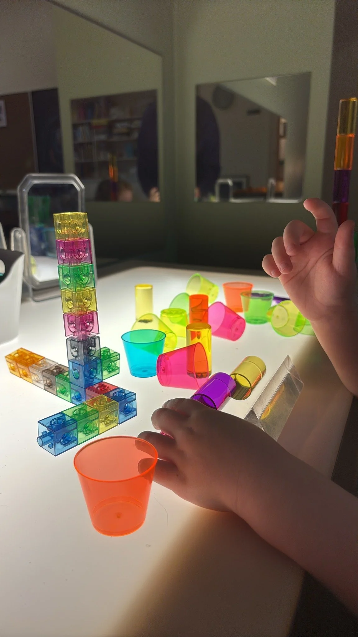 Child at Youngset Preschool building a colorful structure with translucent plastic cups and blocks on a light table, with a mirror behind and a person in the reflection.
