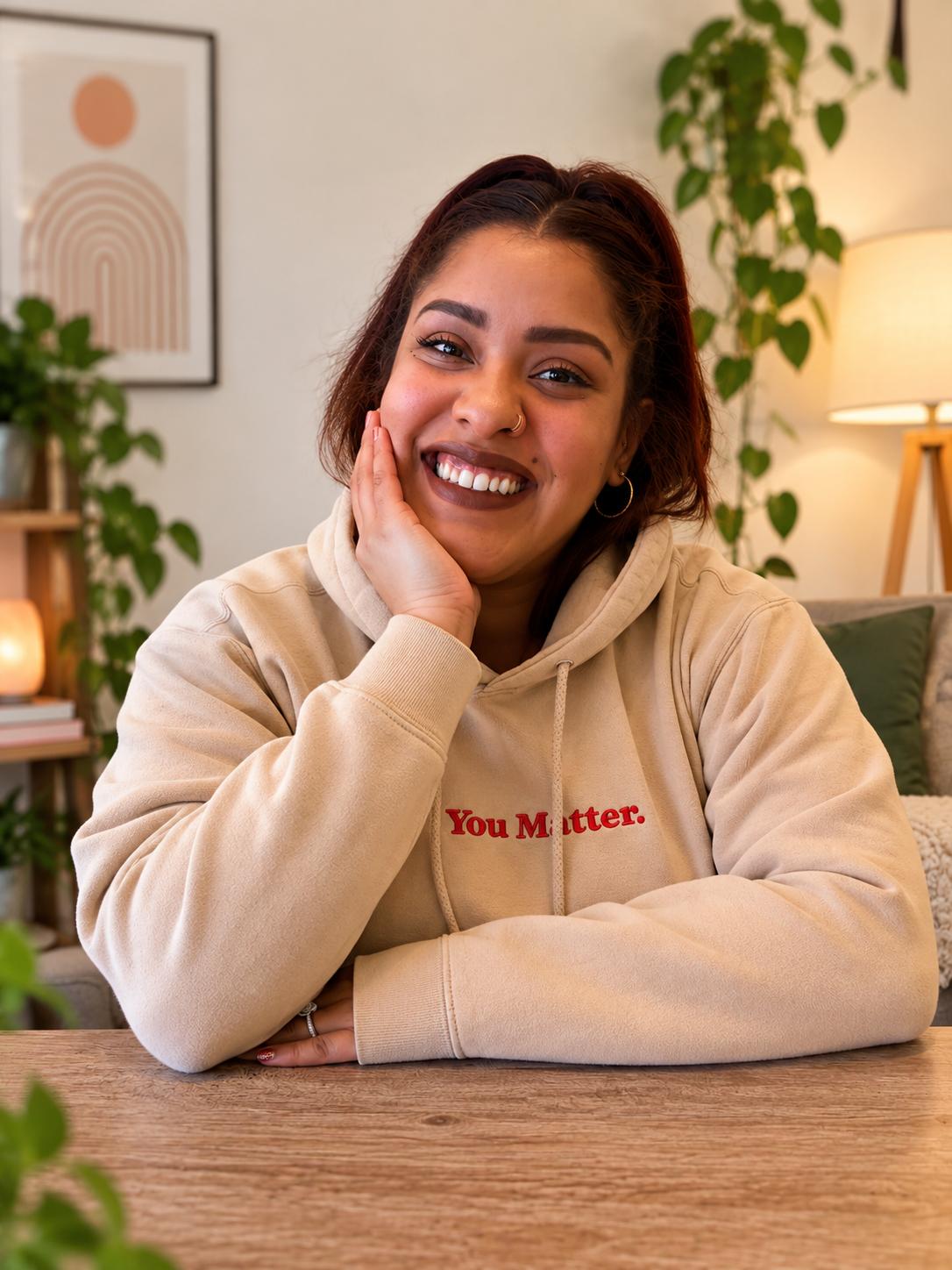 A young woman with dark hair, wearing a beige hoodie with red text, smiling, sitting at a wooden table in a cozy room with green plants and a framed artwork in the background.