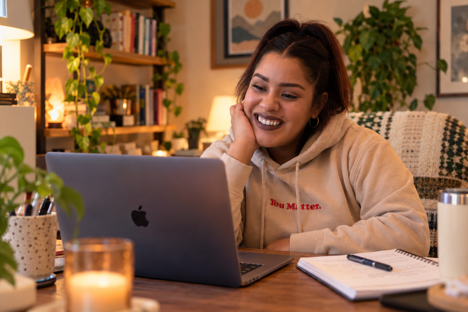 A woman with brown hair, wearing earrings and a beige hoodie with 'You Matter.' written on it, smiling at a laptop in a cozy indoor setting with warm lighting, plants, books, and decorative items.