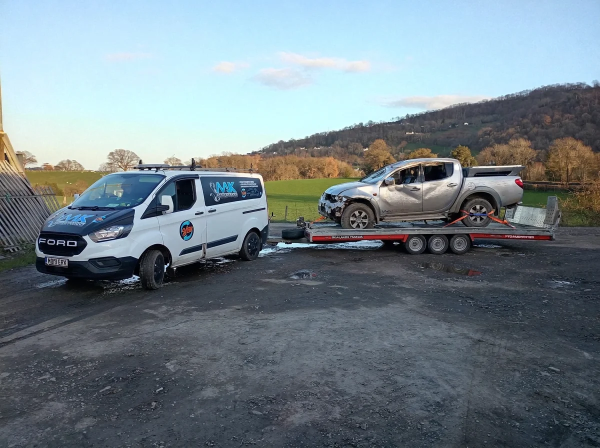 A white service van with 'NX' and other stickers parked next to a flatbed trailer carrying a damaged silver pickup truck in an open rural area with hills and trees in the background.