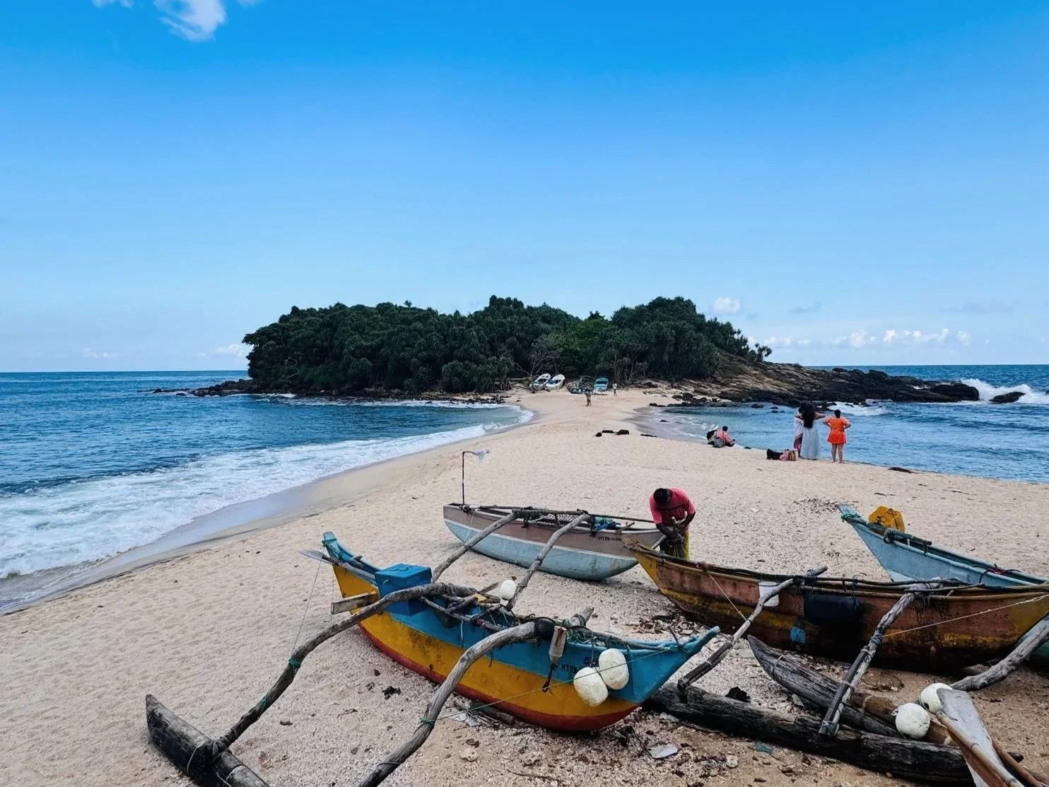 Colourful fishing boats on the sand beside Blue Beach Island in Nilwella, with a green island across the water.