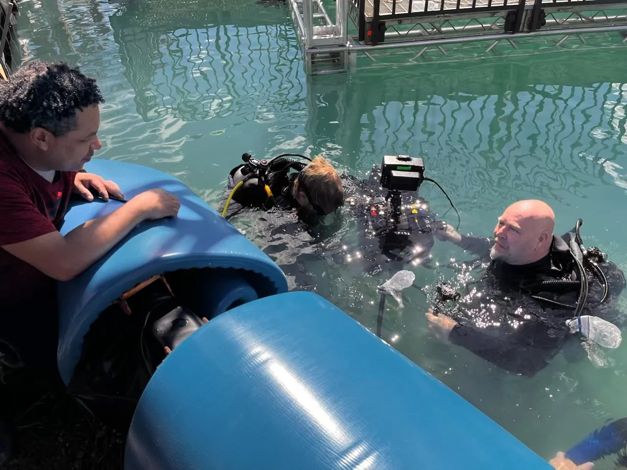 Two divers in a pool with scuba gear, one is submerged and working with underwater equipment while another observes in the water; a person on the pool deck watches them.