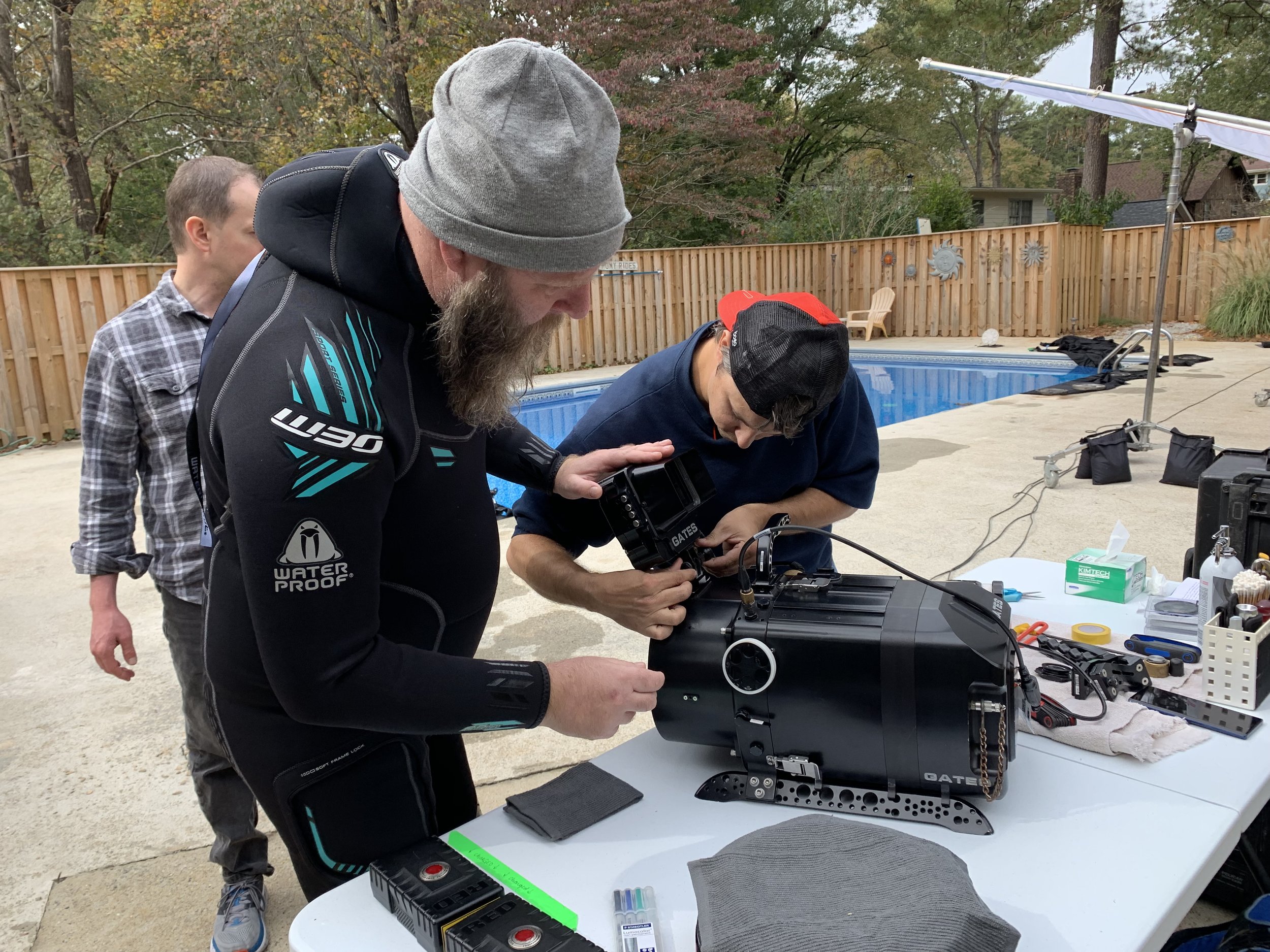 Several men working on electronic equipment outdoors near a swimming pool, with tools and devices on a table.