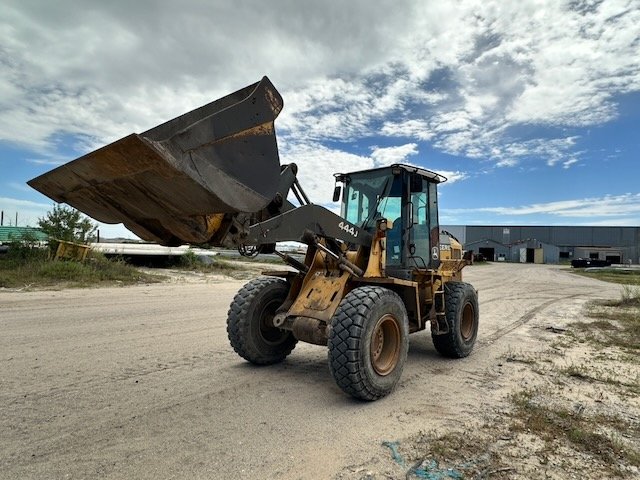 Construction yellow front loader with a large bucket raised, parked on a dirt lot under a partly cloudy sky.