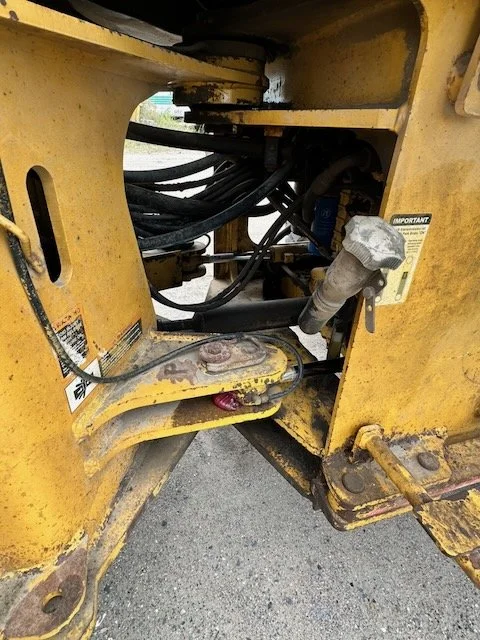 Close-up of the internal mechanical components of a yellow construction machine, showing hydraulic hoses, metal parts, and labels.