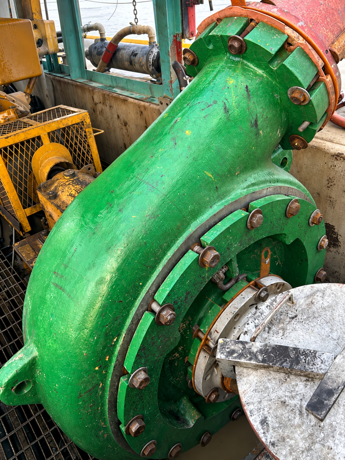 Close-up of large green industrial valve with bolts, attached to piping and machinery, located outdoors near water.
