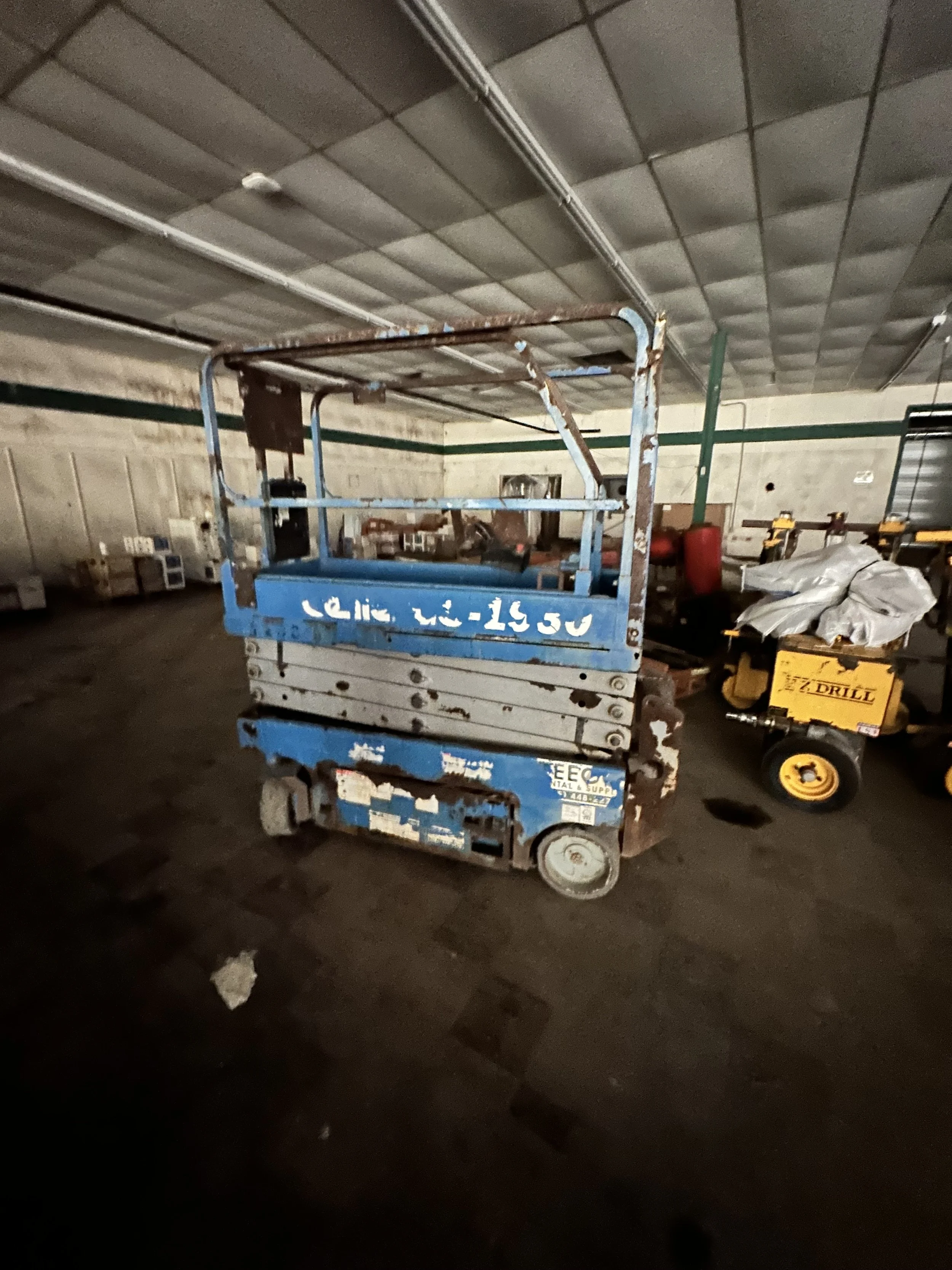 Blue industrial scissor lift parked inside a dim warehouse beneath exposed metal roofing, with a yellow floor drill nearby.
