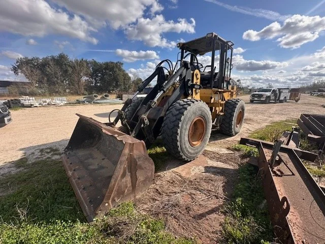 A yellow bulldozer with large black tires parked on dirt and grass at an outdoor construction or storage yard, with a partly cloudy sky above.