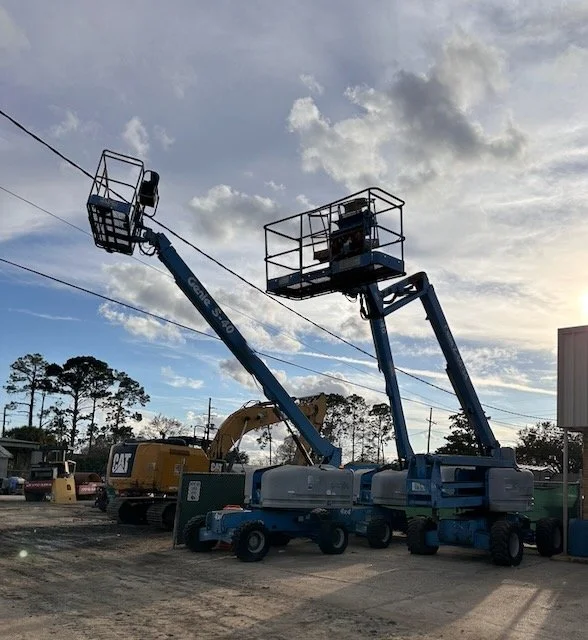 Construction equipment yard with two blue boom lifts, a CAT excavator, utility lines, and a cloudy sky