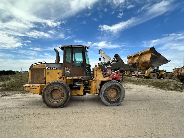 A yellow John Deere front loader construction vehicle with a scoop attachment, parked on a gravel surface under a partly cloudy sky.