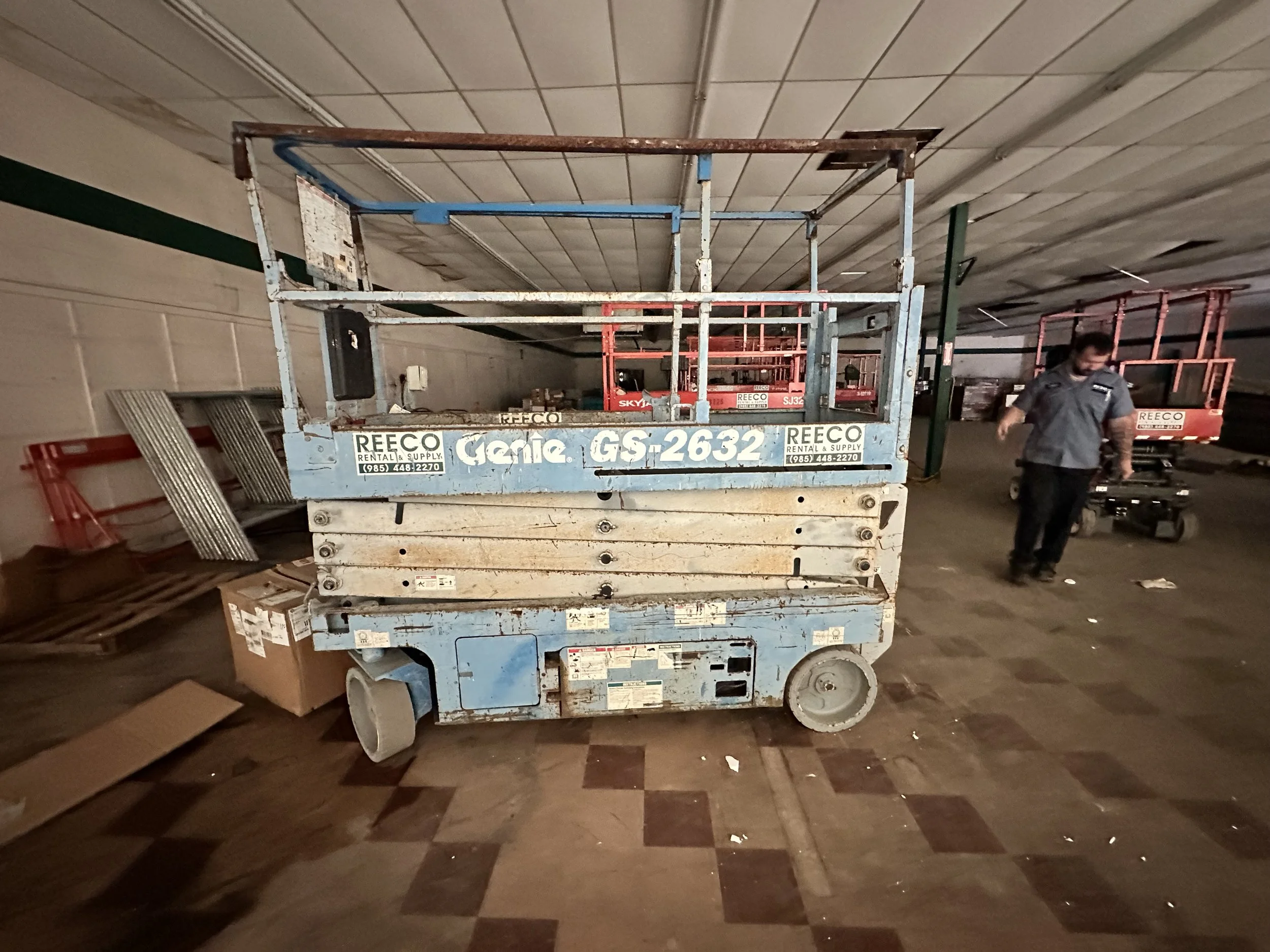 Blue Genie GS-2632 scissor lift in a warehouse, with additional lifts and a worker in the background.