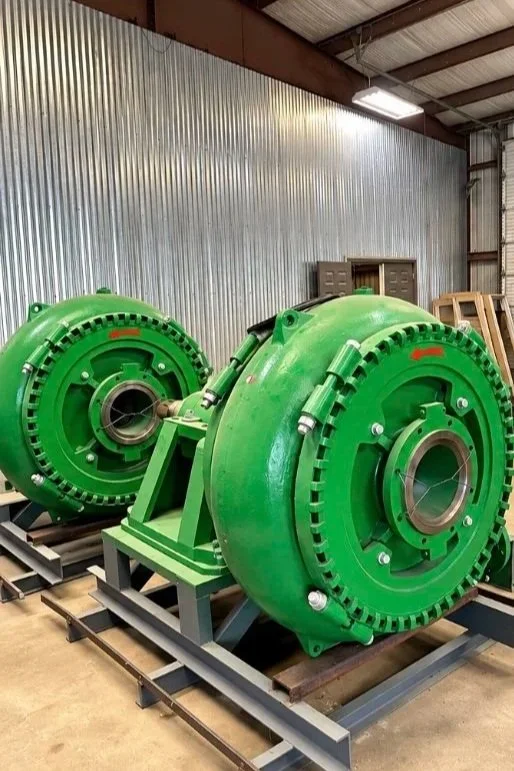 Two large, green industrial pumps or motors mounted on metal stands inside a warehouse with corrugated metal walls and a high ceiling.