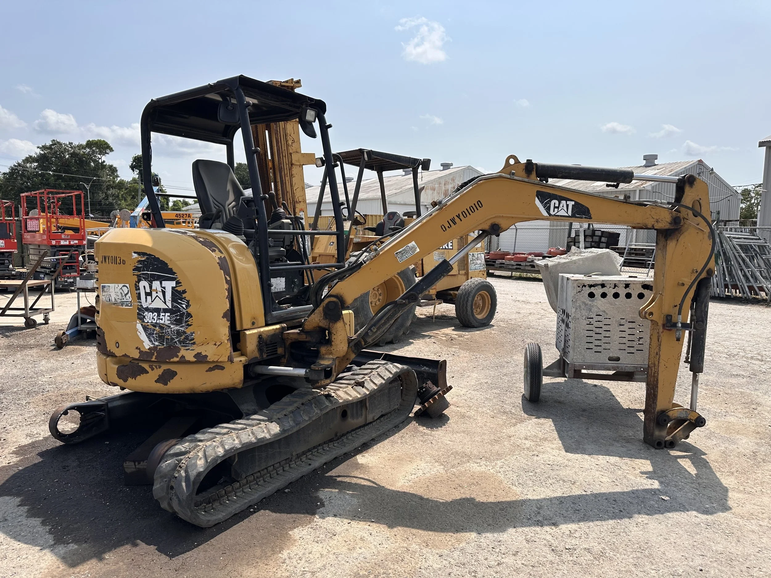 Used yellow CAT compact excavator parked at a construction equipment yard, with other heavy machinery and industrial buildings in the background