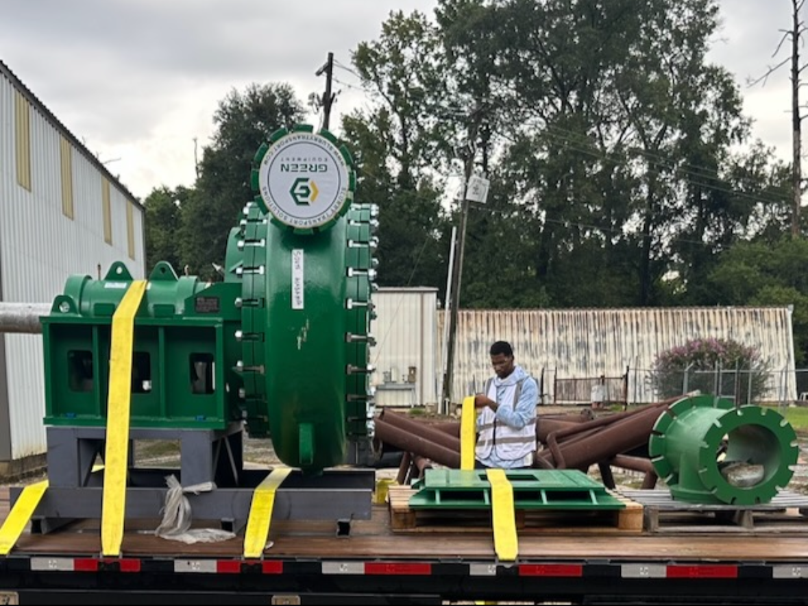 A man inspecting large, industrial green machinery parts on a flatbed truck outdoors on a cloudy day.