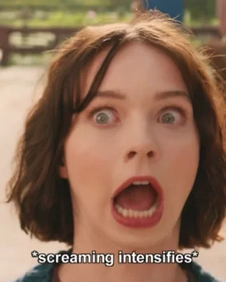 Close-up of a woman with short brown hair and surprised facial expression, mouth open, eyes wide, outdoors with a fence and trees in the background.